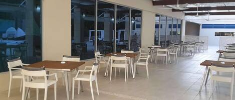 Een ruime eethoek binnen met meerdere tafels en stoelen, grote ramen en plafondventilatoren.