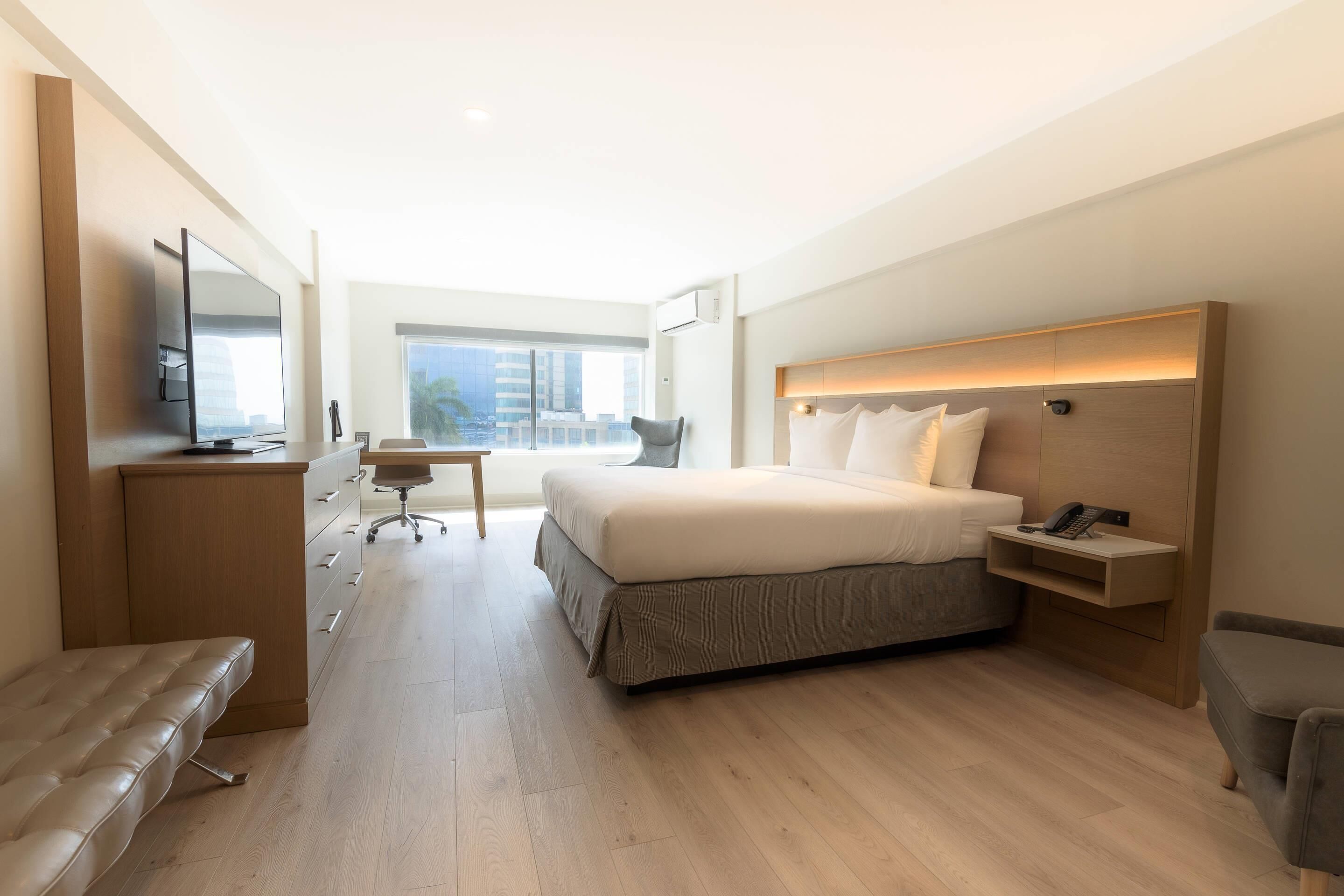 A modern hotel room with a large bed, a desk, a chair, a television, and a view of the city.