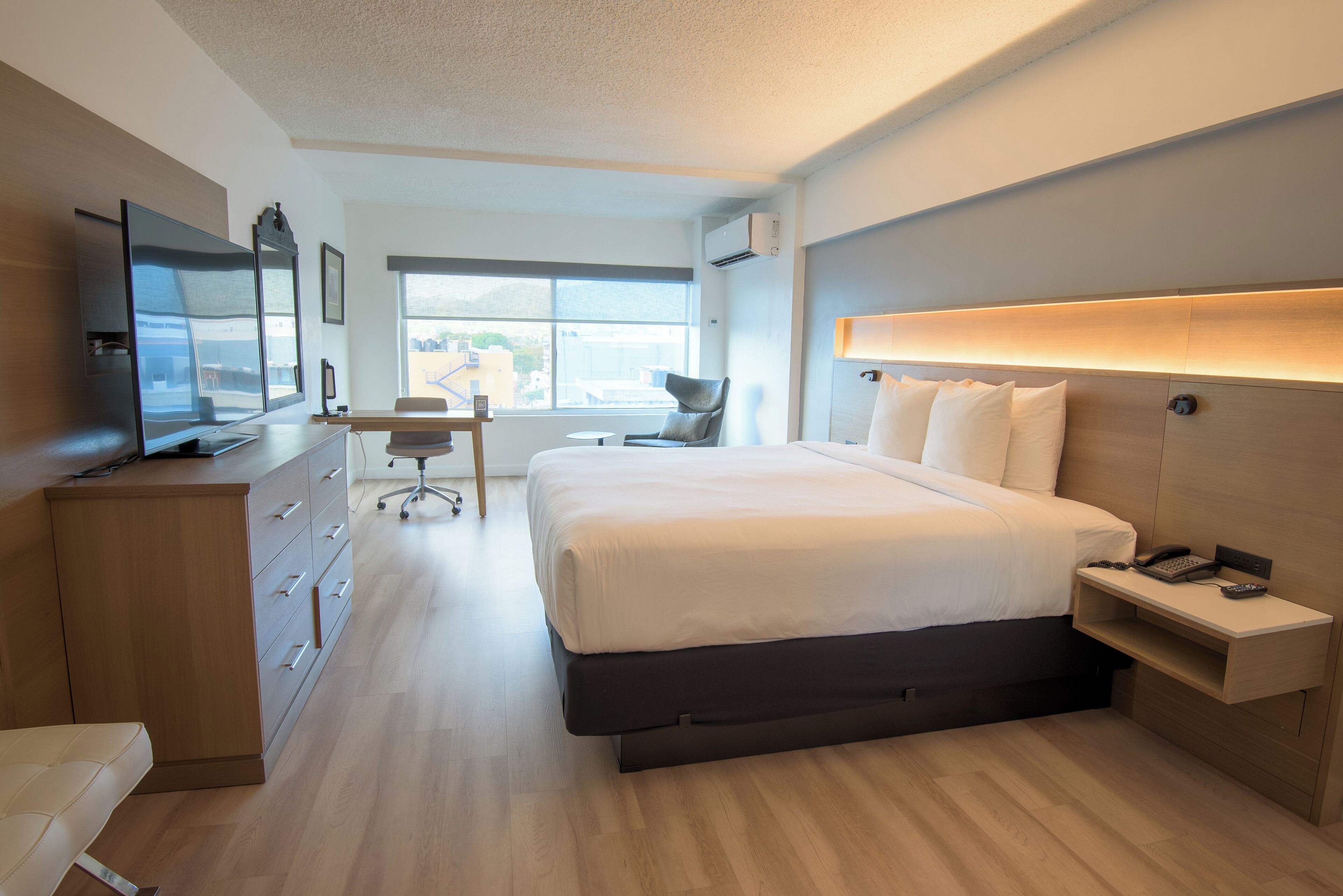 A modern hotel room with a large bed, a desk, a television, and a view of the city through the window.