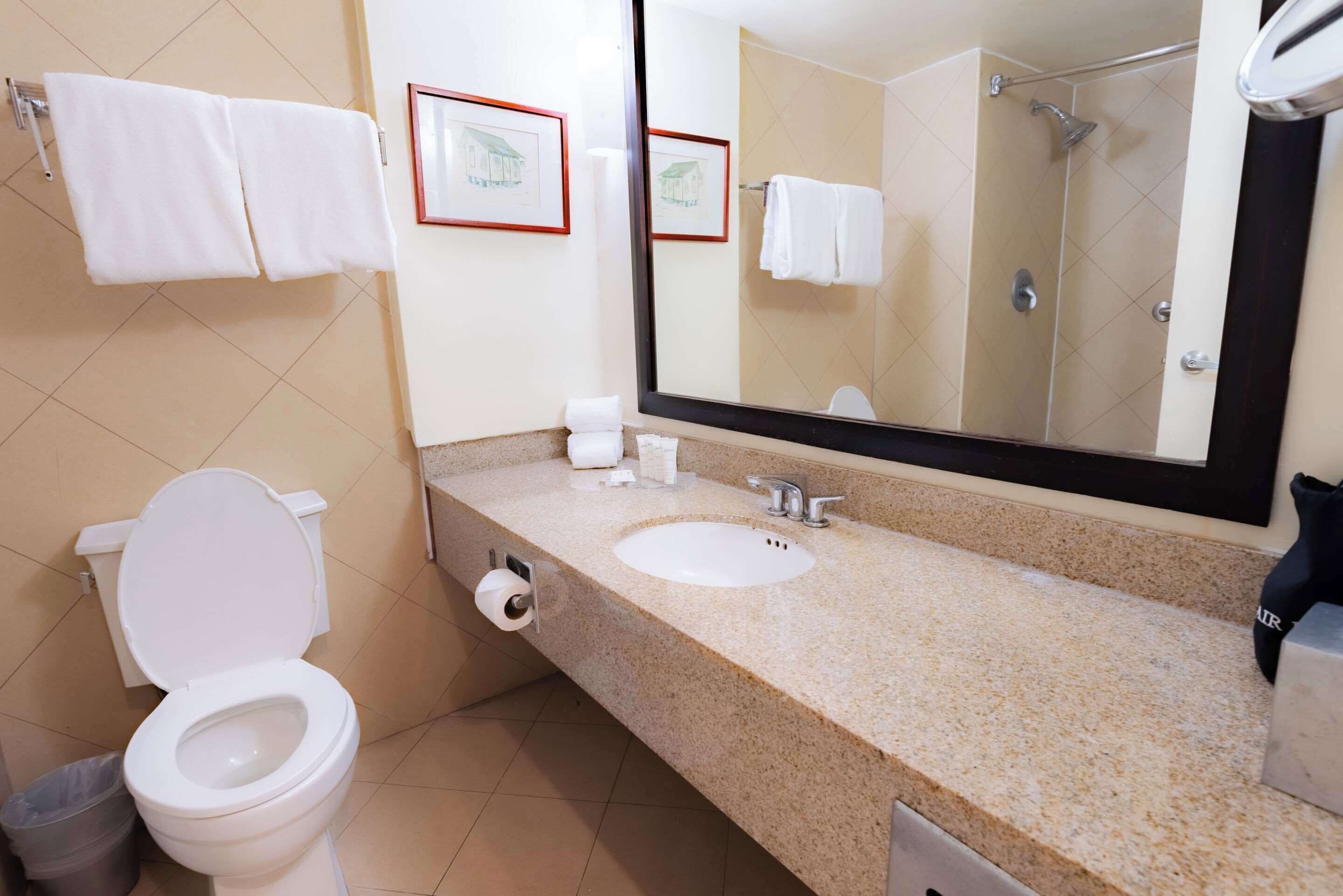 A bathroom with a toilet, sink, and mirror. There are towels hanging on the wall and a trash can.