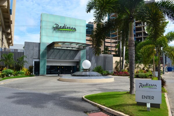 A Radisson hotel entrance with a modern glass facade, a spherical water feature, and a sign indicating the hotel is in Trinidad.