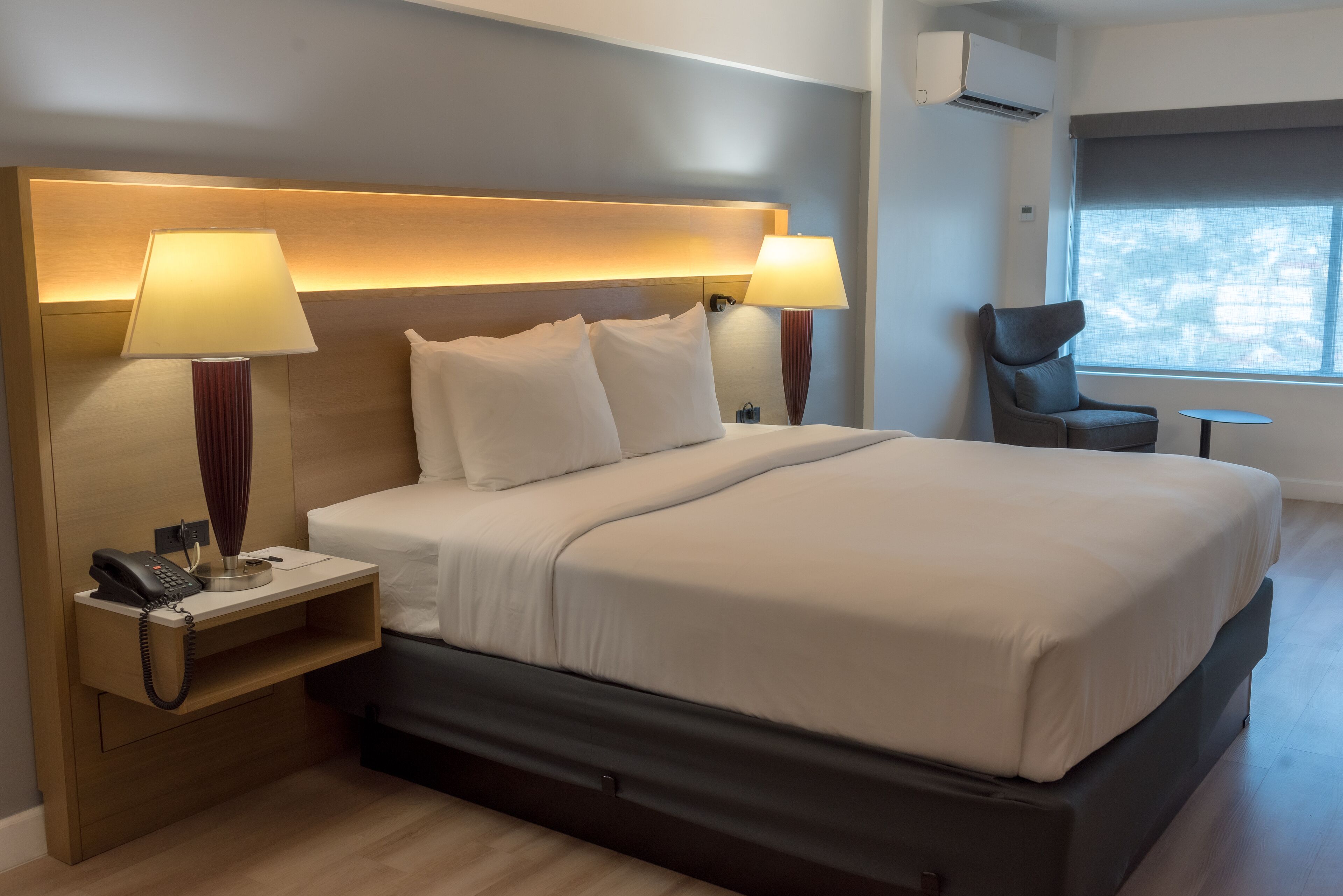 A modern hotel room with a large bed, two bedside lamps, a chair, a small table, and a phone on the nightstand.