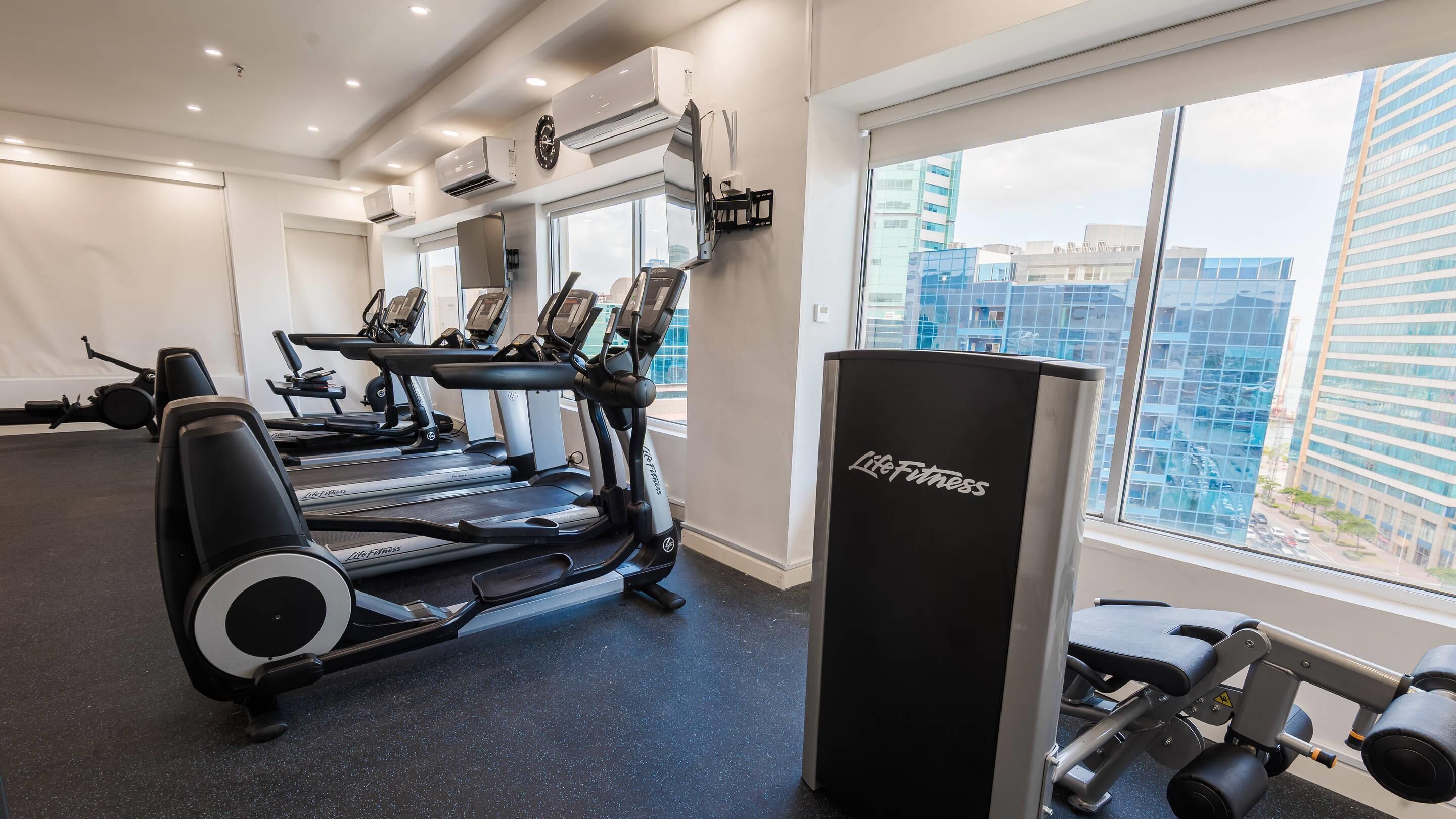 Ett gym med löpband, en viktbänk och ett stort fönster med utsikt över staden.