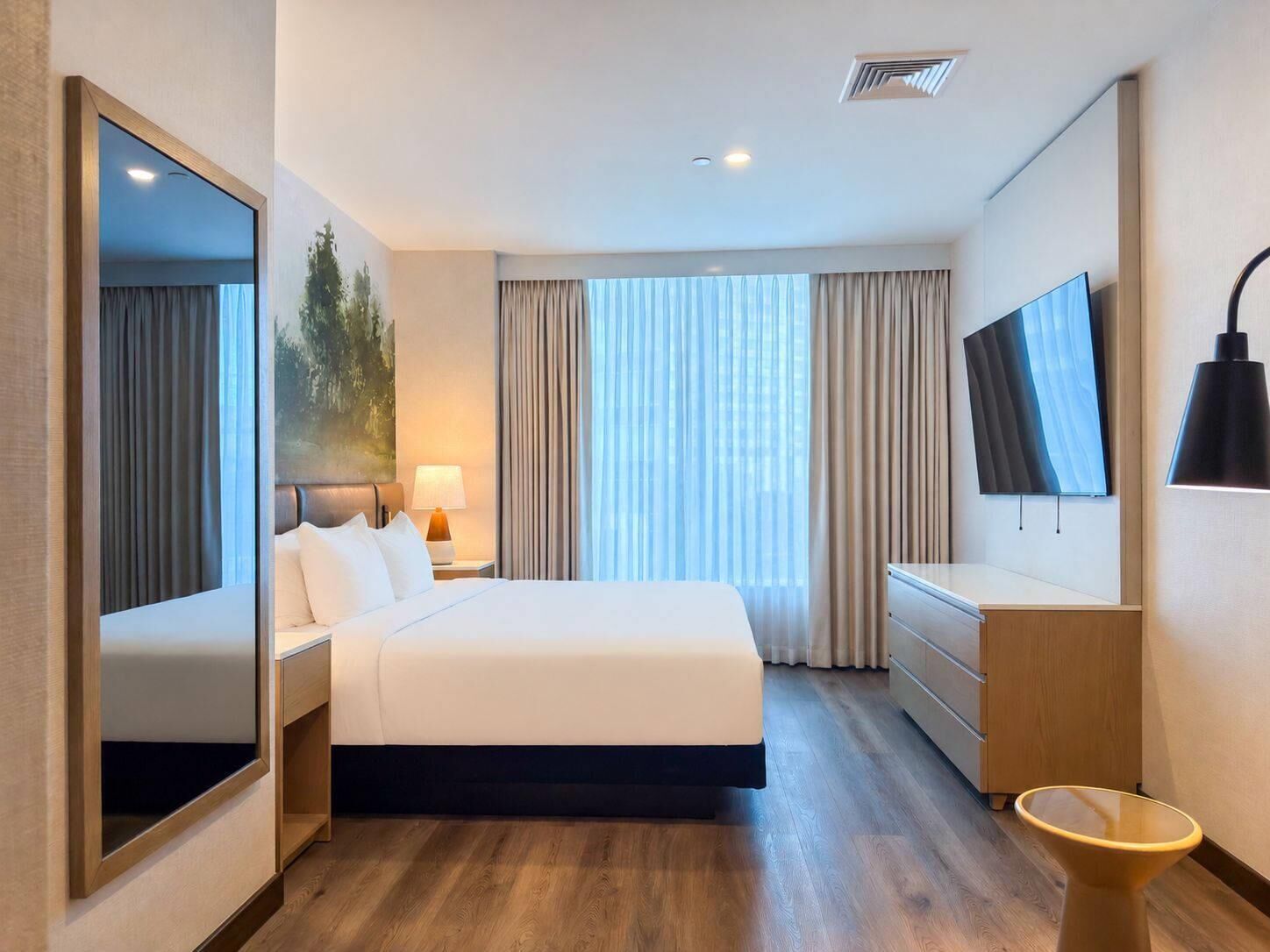 Habitación de hotel con cama, televisor, escritorio, silla, lámpara y un ventanal grande con cortinas.