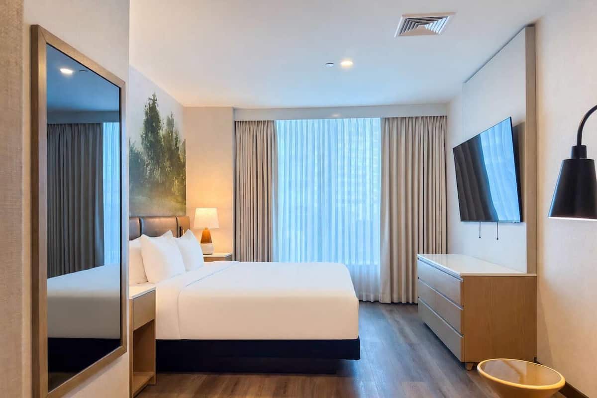 A hotel room with a bed, a TV, a desk, a chair, a lamp, and a large window with curtains.
