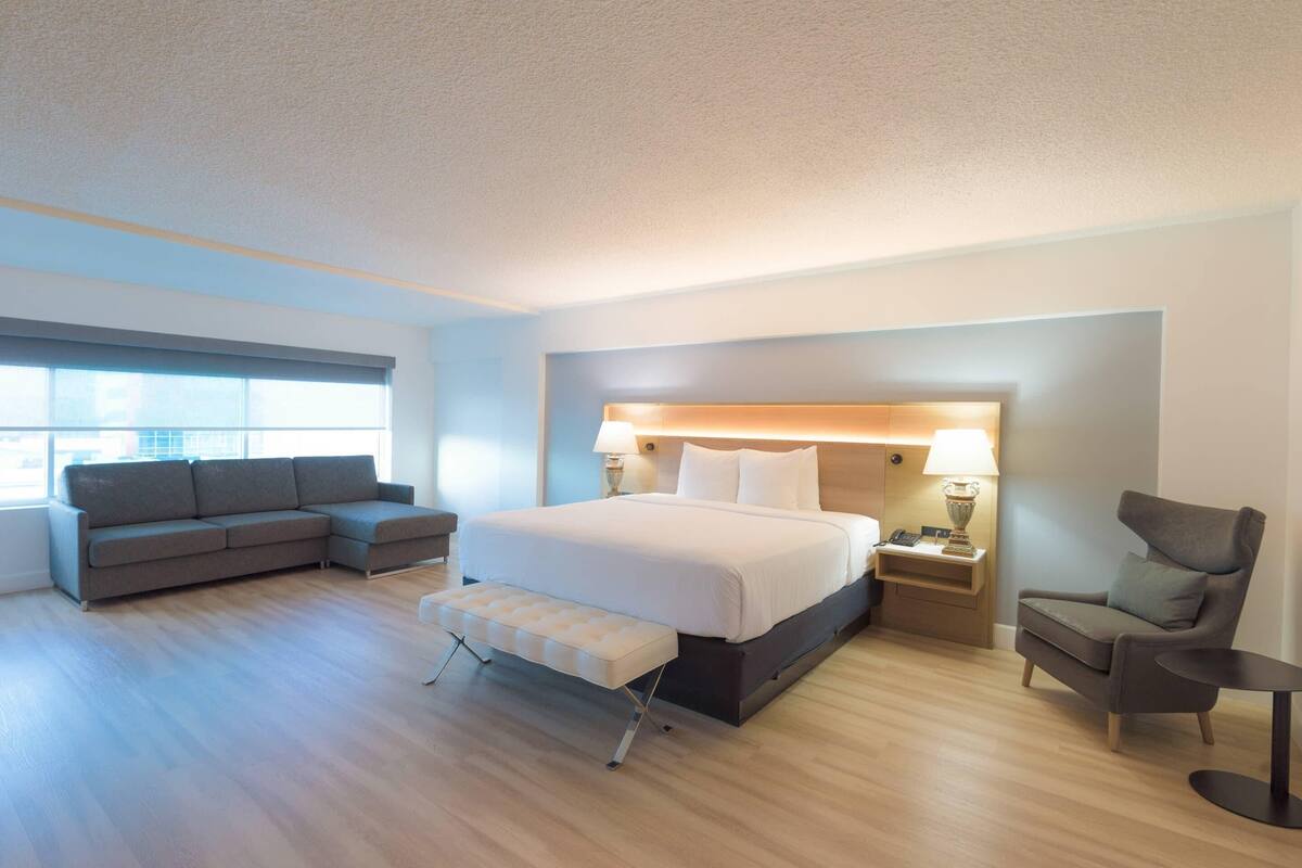 A modern hotel room with a large bed, a sofa, a chair, a small table, and a nightstand.