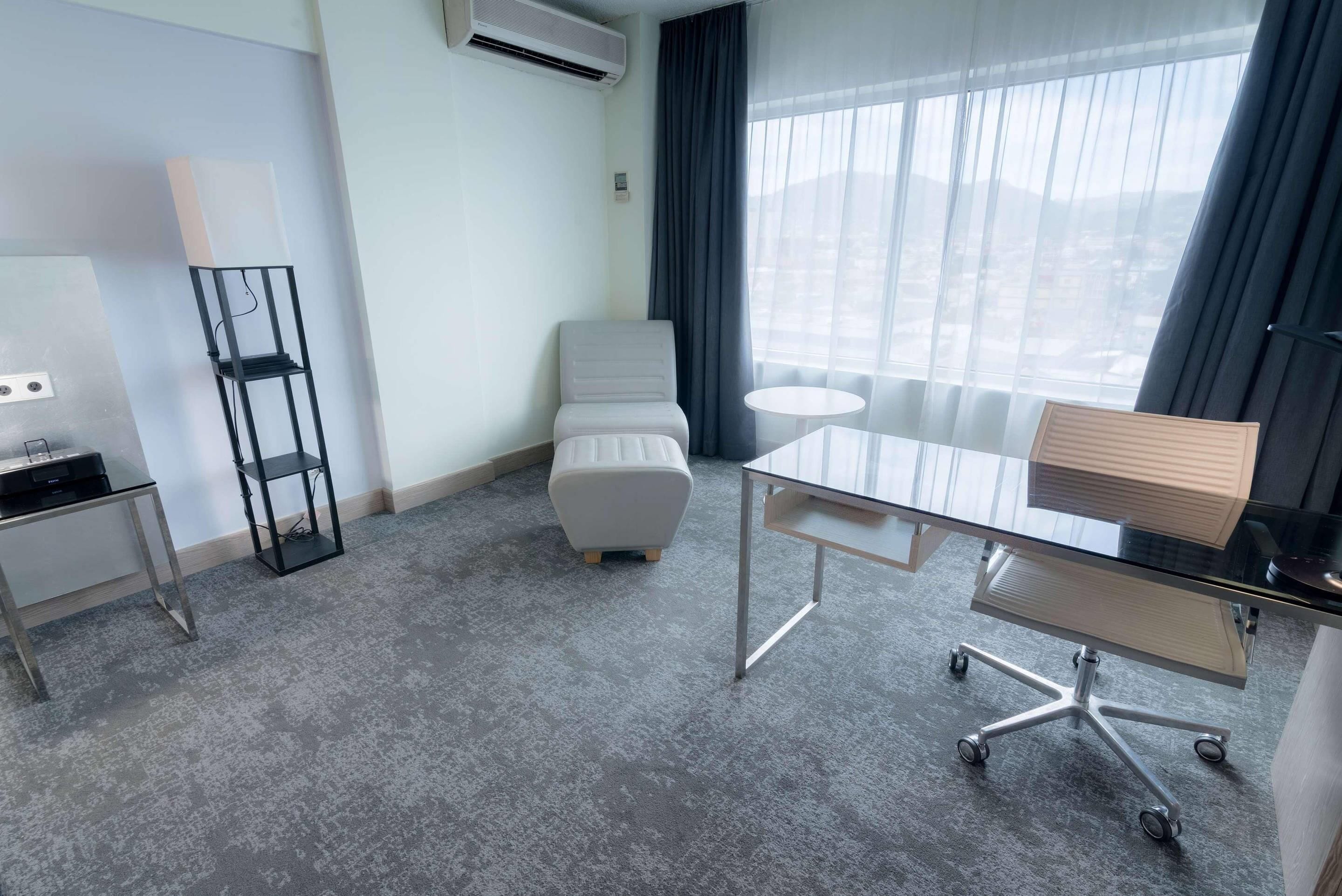 Une chambre d’hôtel avec une grande fenêtre, une chaise blanche, une table en verre et un petit bureau avec une lampe.