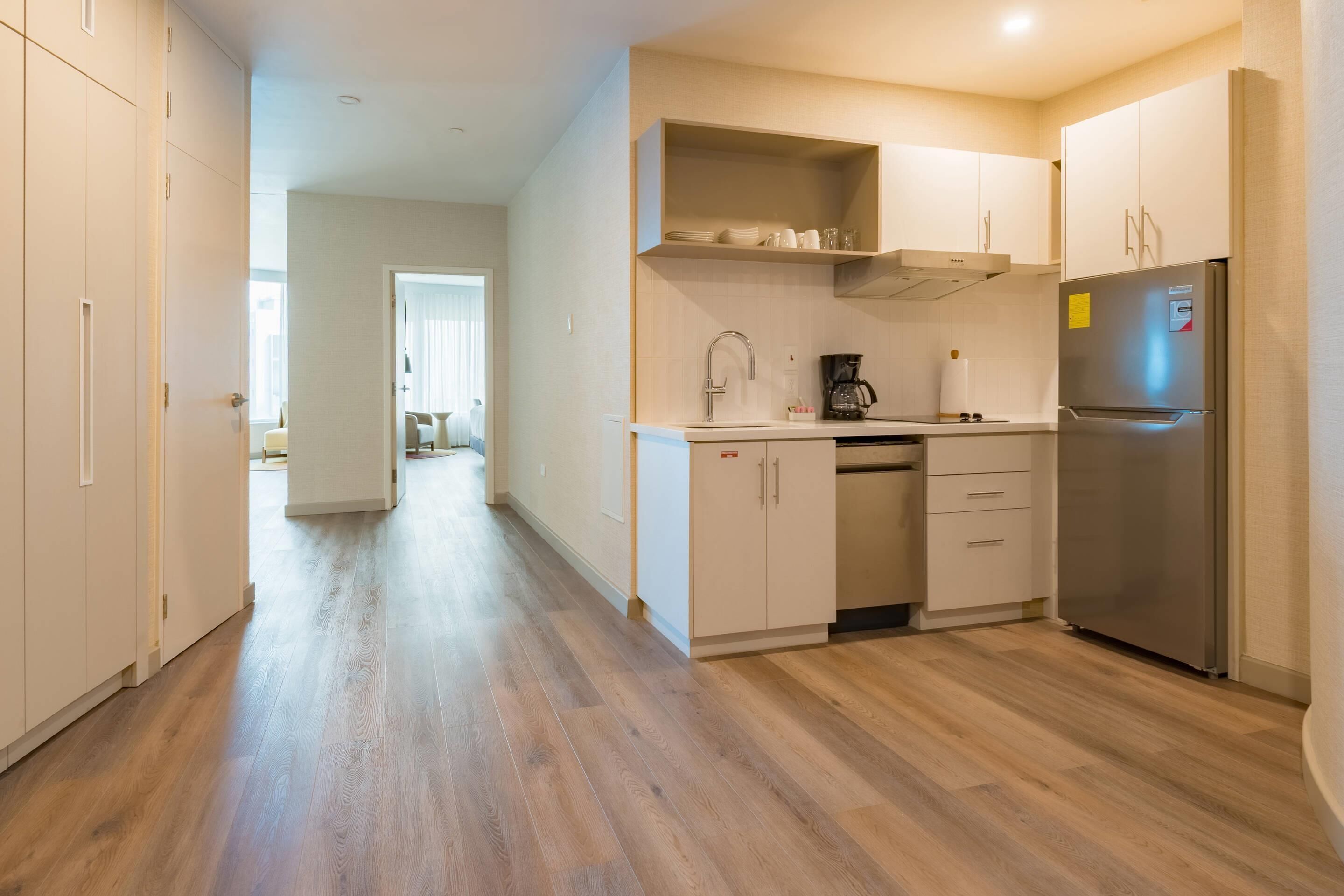 Una cocina moderna con suelo de madera, armarios blancos, un refrigerador de acero inoxidable y una cafetera.