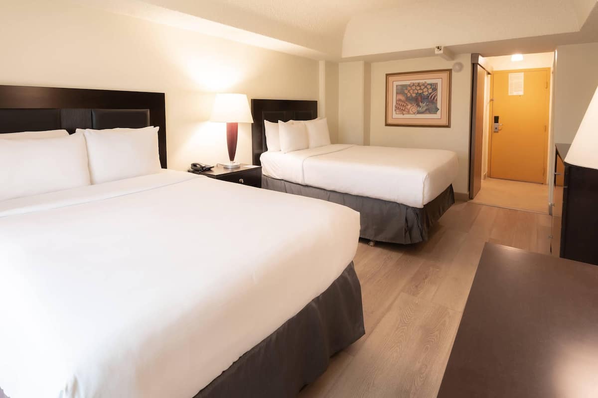 A hotel room with two beds, a desk, a nightstand, a lamp, and a door leading to another room.