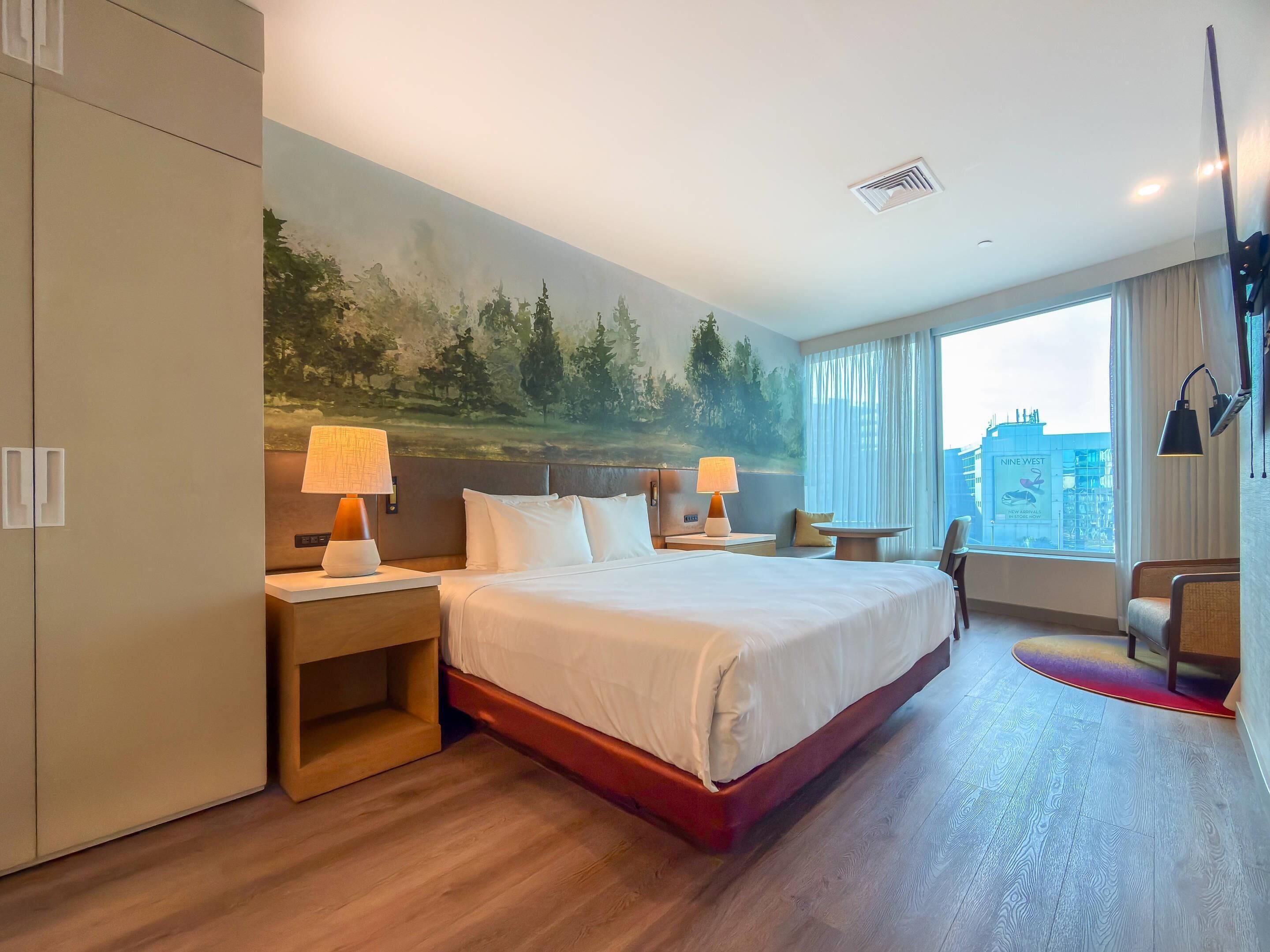 A hotel room with a large bed, two bedside tables with lamps, a chair, a small table, and a painting on the wall.