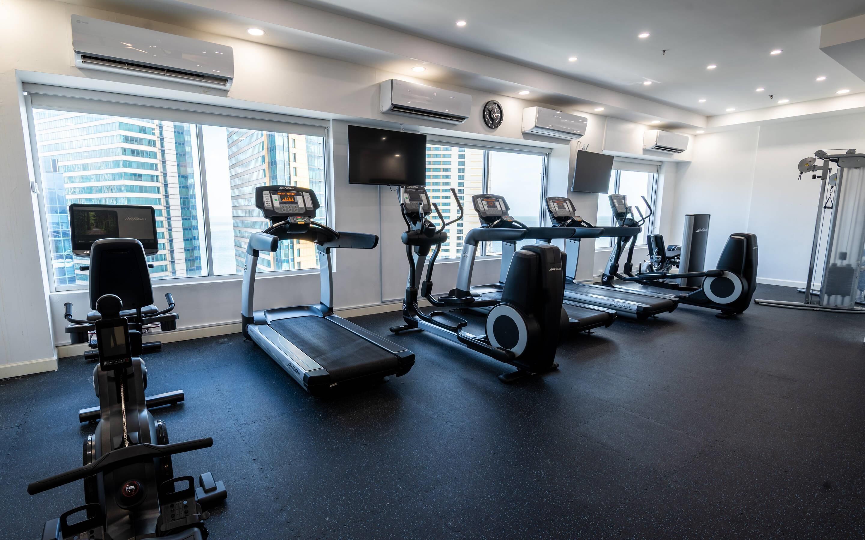 A gym with treadmills, ellipticals, and a stationary bike, large windows, and air conditioning units.