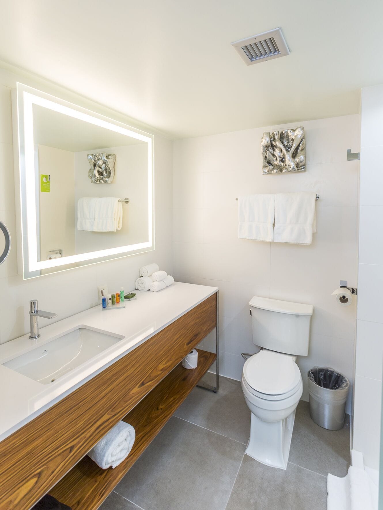 A modern bathroom with a large mirror, white sink, wooden vanity, toilet, and a trash can.
