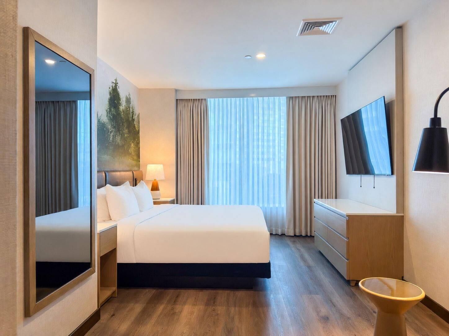 Quarto de hotel com cama, televisão, escrivaninha, cadeira, espelho e janela com cortinas.
