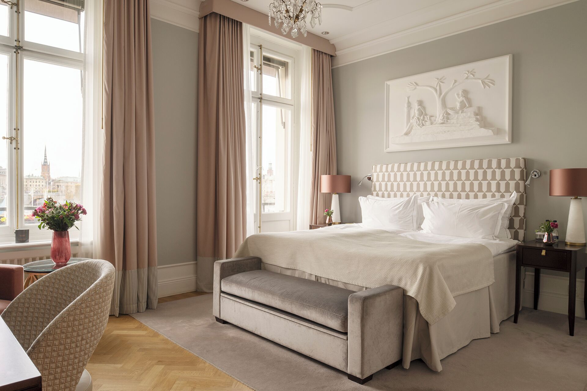 A bedroom with a large bed, a bedside table, a chair, and a view of the city through the window.