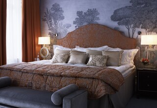 A bedroom with a large bed, two bedside tables with lamps, a headboard with a floral pattern, and a wall mural of trees.