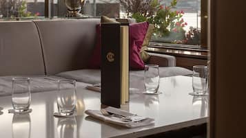 A hotel room with a dining table set for two, a sofa, a lamp, and a view of the cityscape.