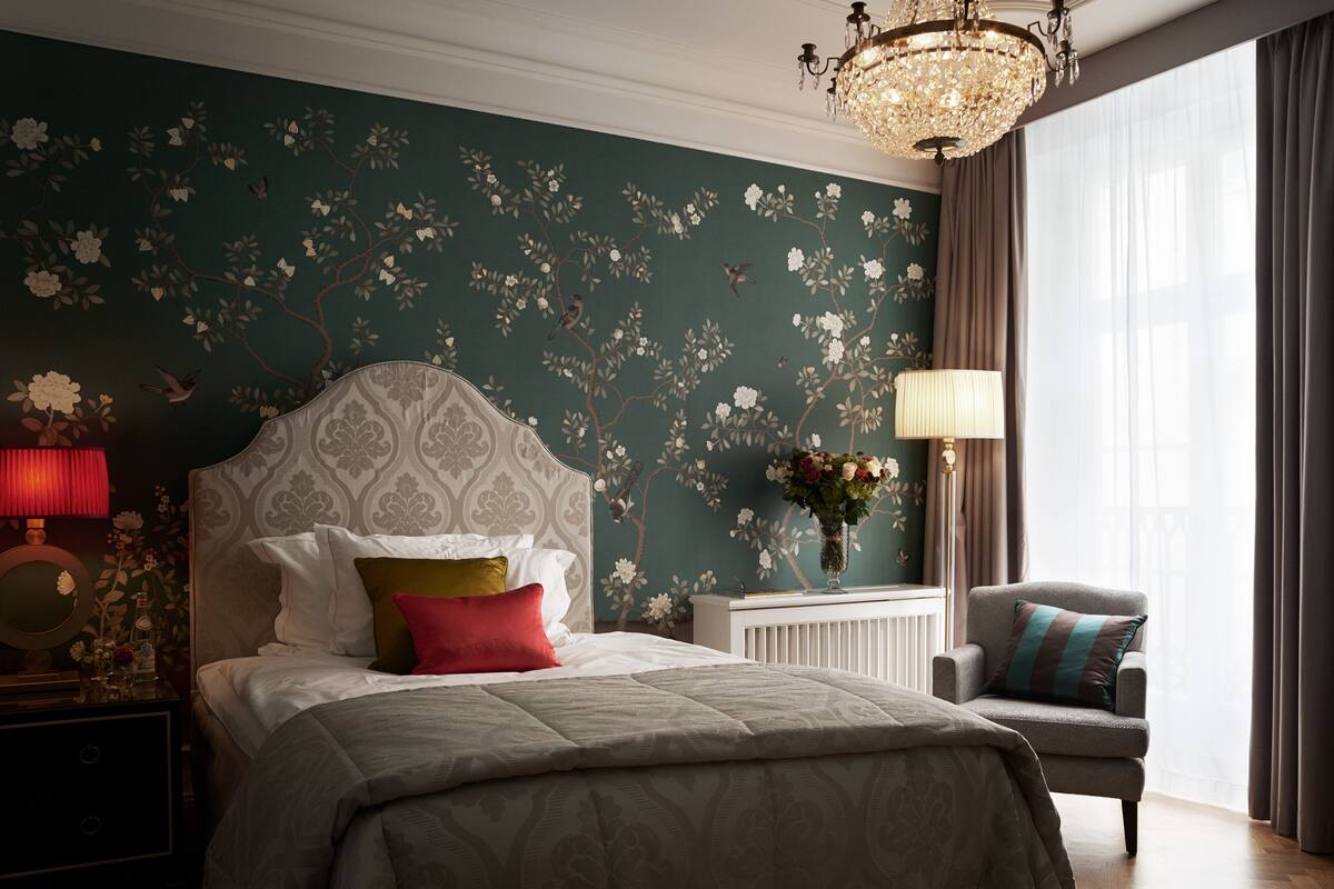 A bedroom with a large bed, a nightstand, a chair, a chandelier, and a window with curtains.