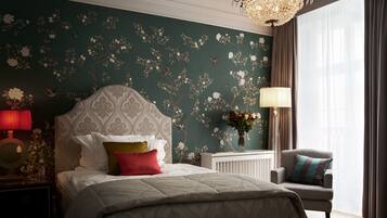 A bedroom with a large bed, a nightstand, a chair, a chandelier, and a window with curtains.