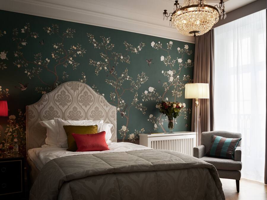 A bedroom with a large bed, a nightstand, a chair, a chandelier, and a window with curtains.