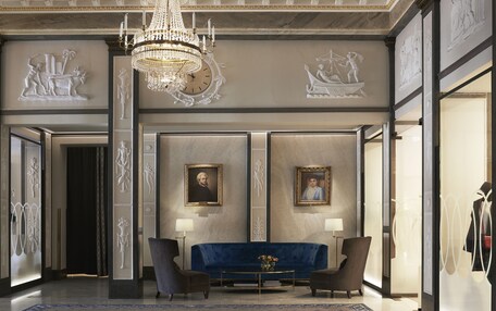 A luxurious lobby with a grand chandelier, marble walls, and classical sculptures. There are two armchairs, a blue sofa, and a coffee table with a vase.