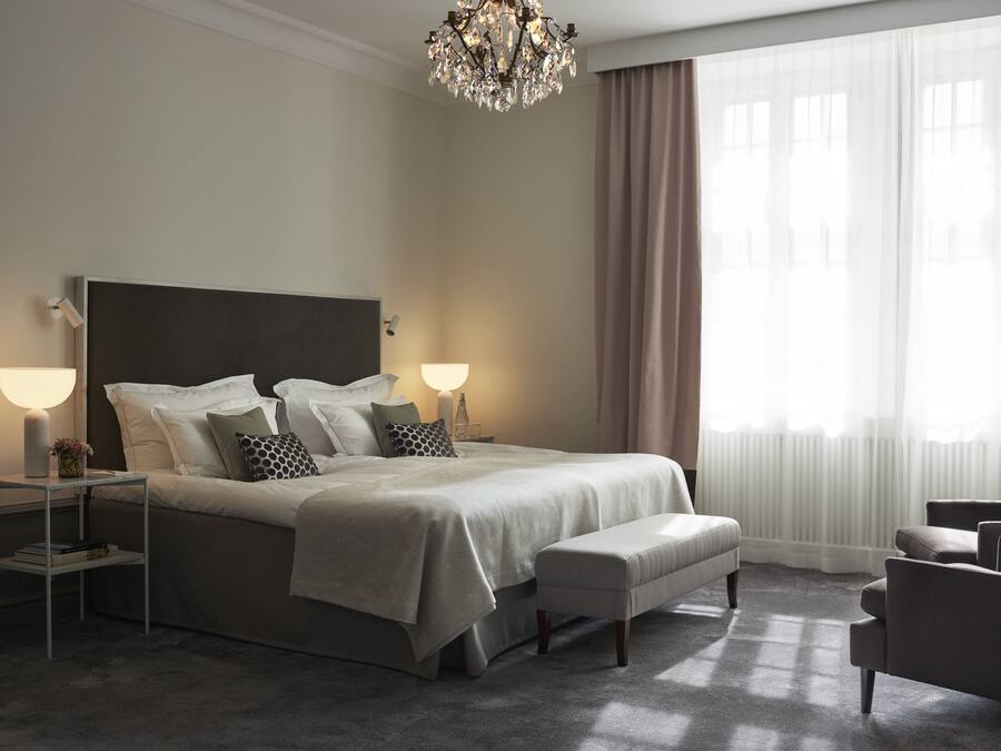 Um quarto bem arrumado com uma cama grande, abajures, um banquinho e uma cadeira.