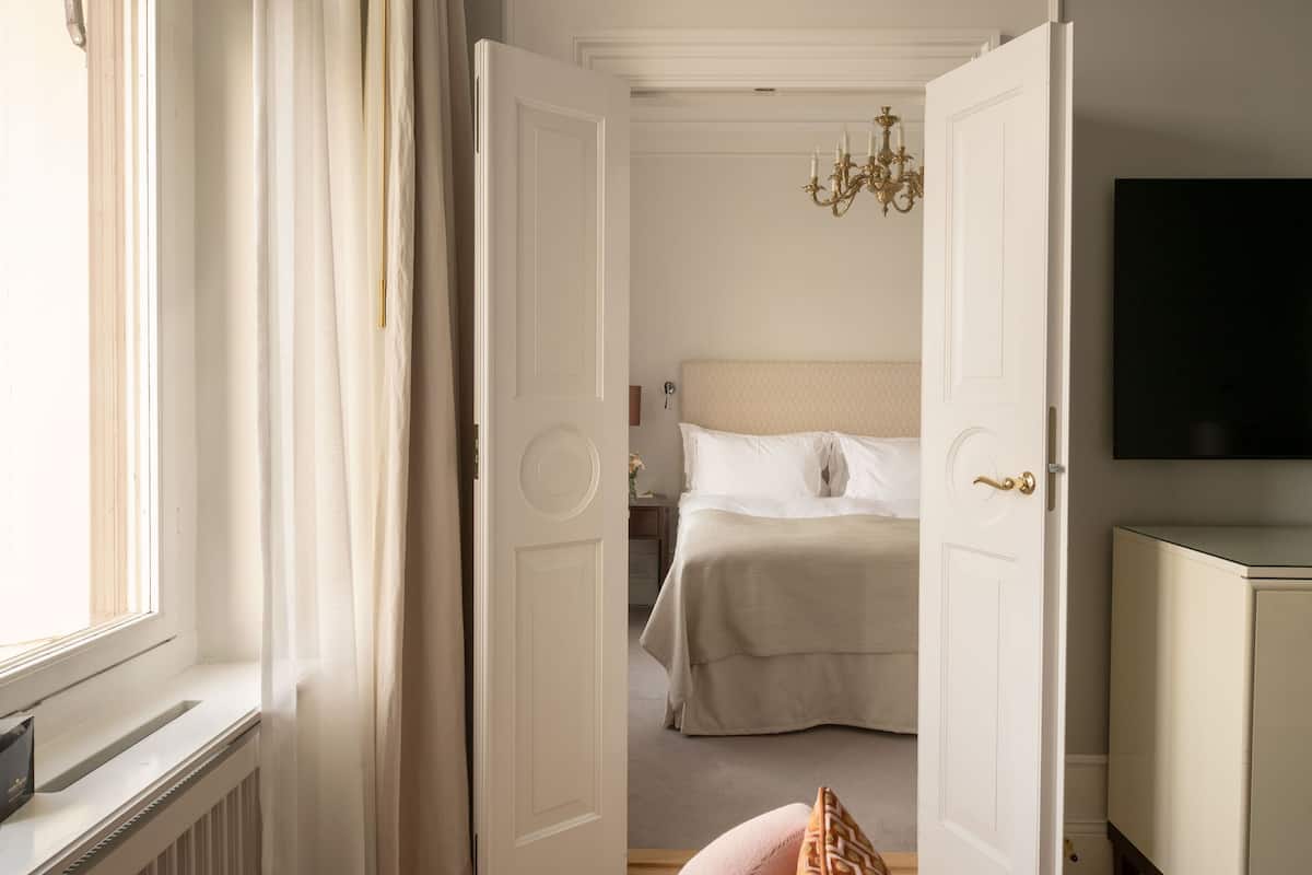 A bedroom with a bed, a nightstand, a dresser, a chandelier, and a window with curtains.