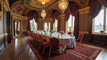Comedor formal con una larga mesa dispuesta para un banquete, candelabros ornamentados y ventanas grandes que dan a vistas de la ciudad.