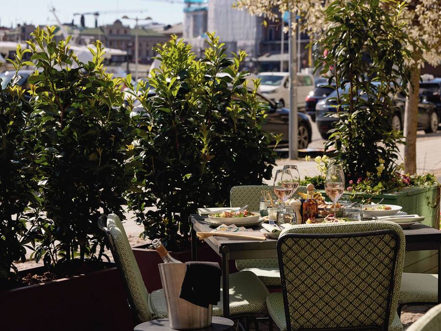 An outdoor dining area with green patterned chairs and tables, surrounded by lush greenery and a cityscape in the background.