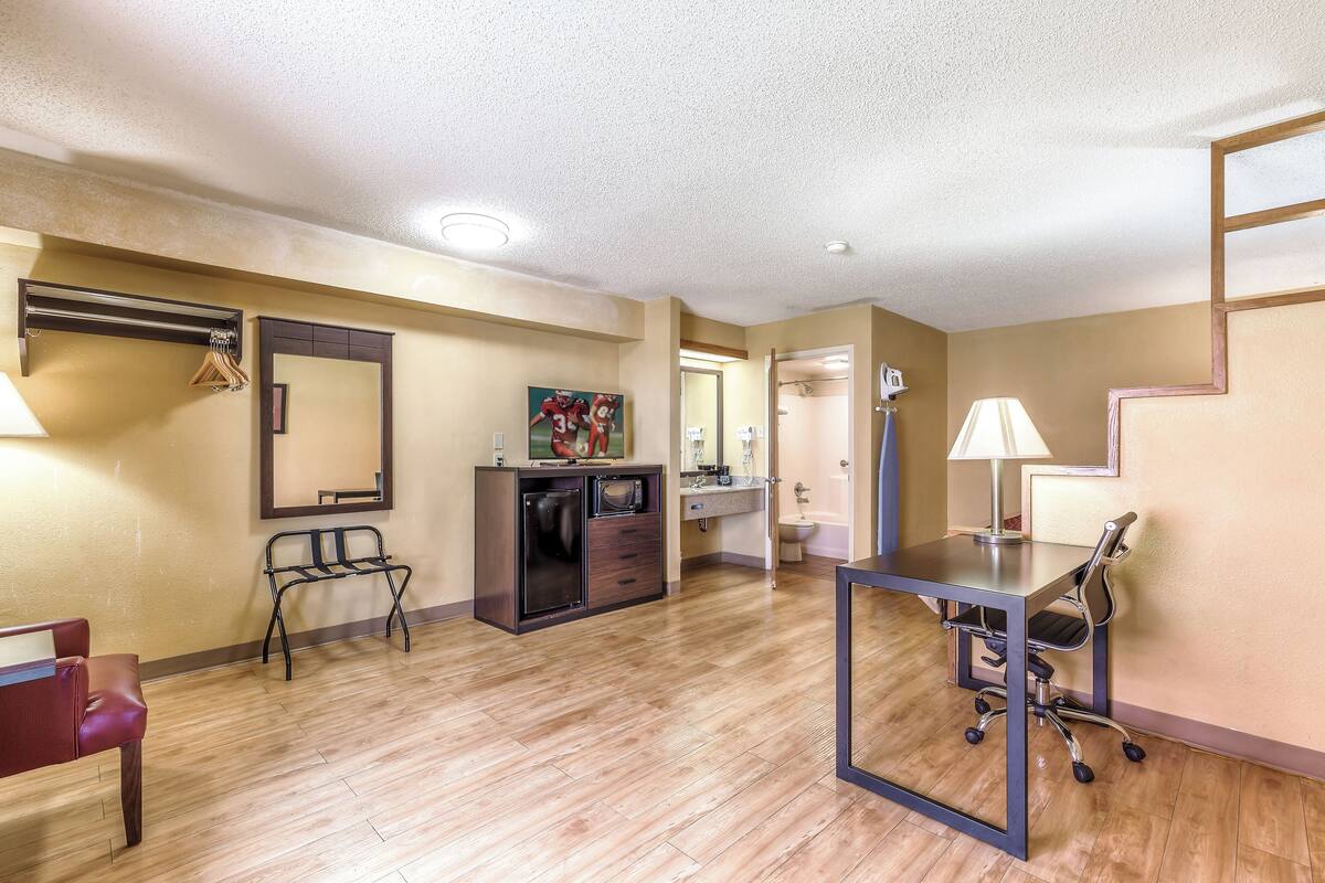 A hotel room with a wooden floor, a desk with a chair, a mirror, a coat rack, and a bathroom visible through an open door.