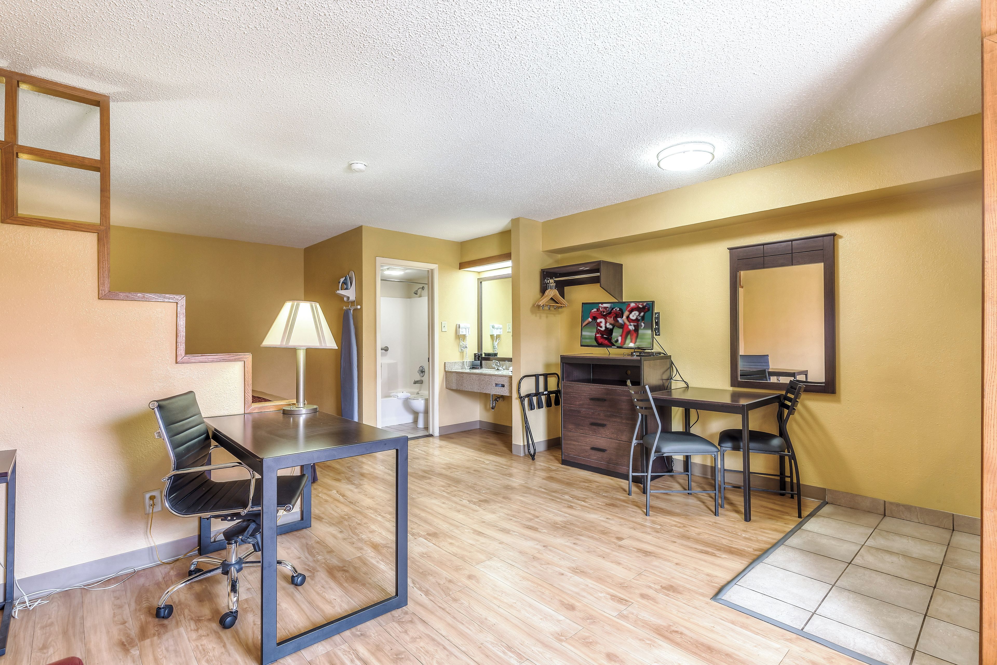 A hotel room with a dining area, a desk with a chair, a TV, a bathroom with a shower, and a staircase.