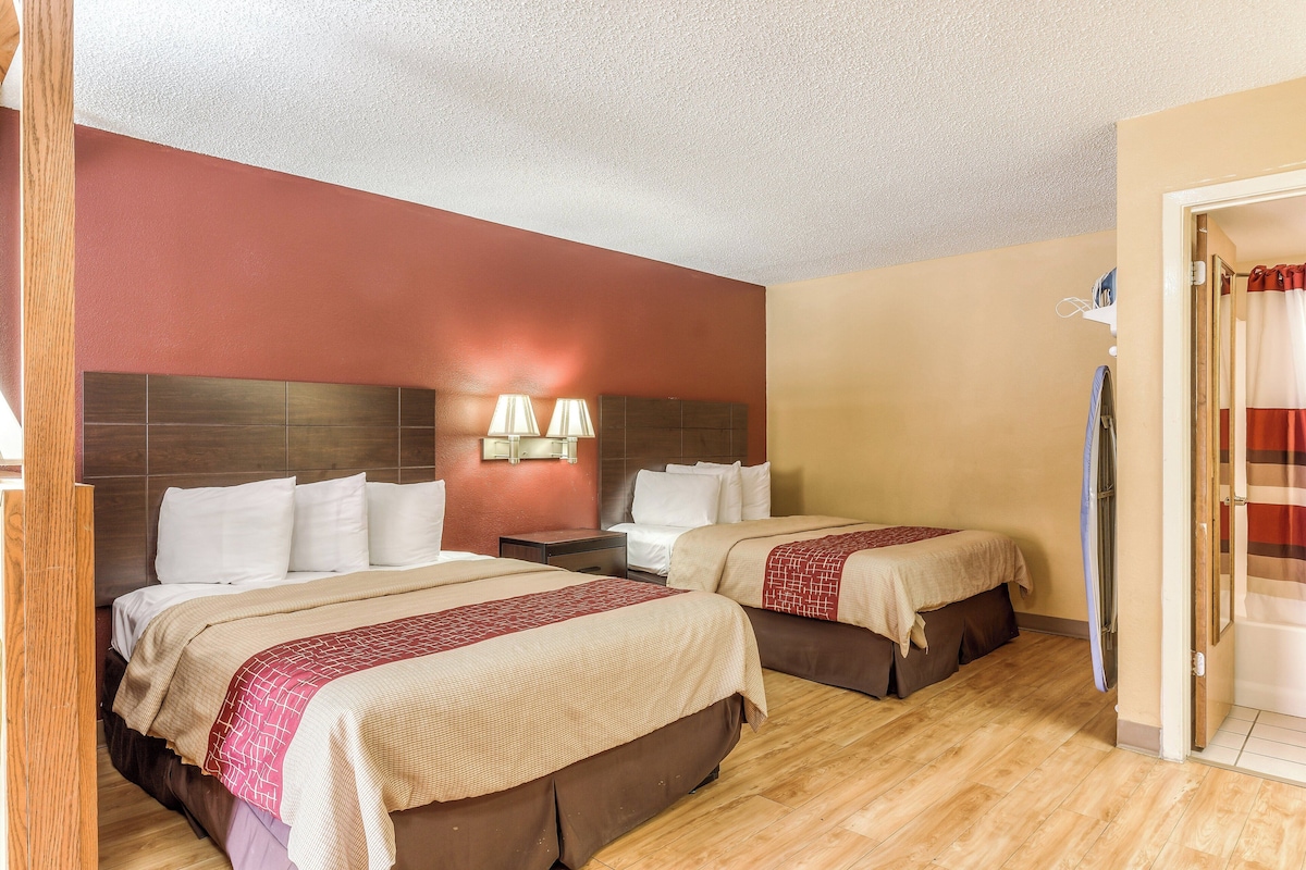 A hotel room with two beds, wooden flooring, a red accent wall, and a bathroom visible through an open door.