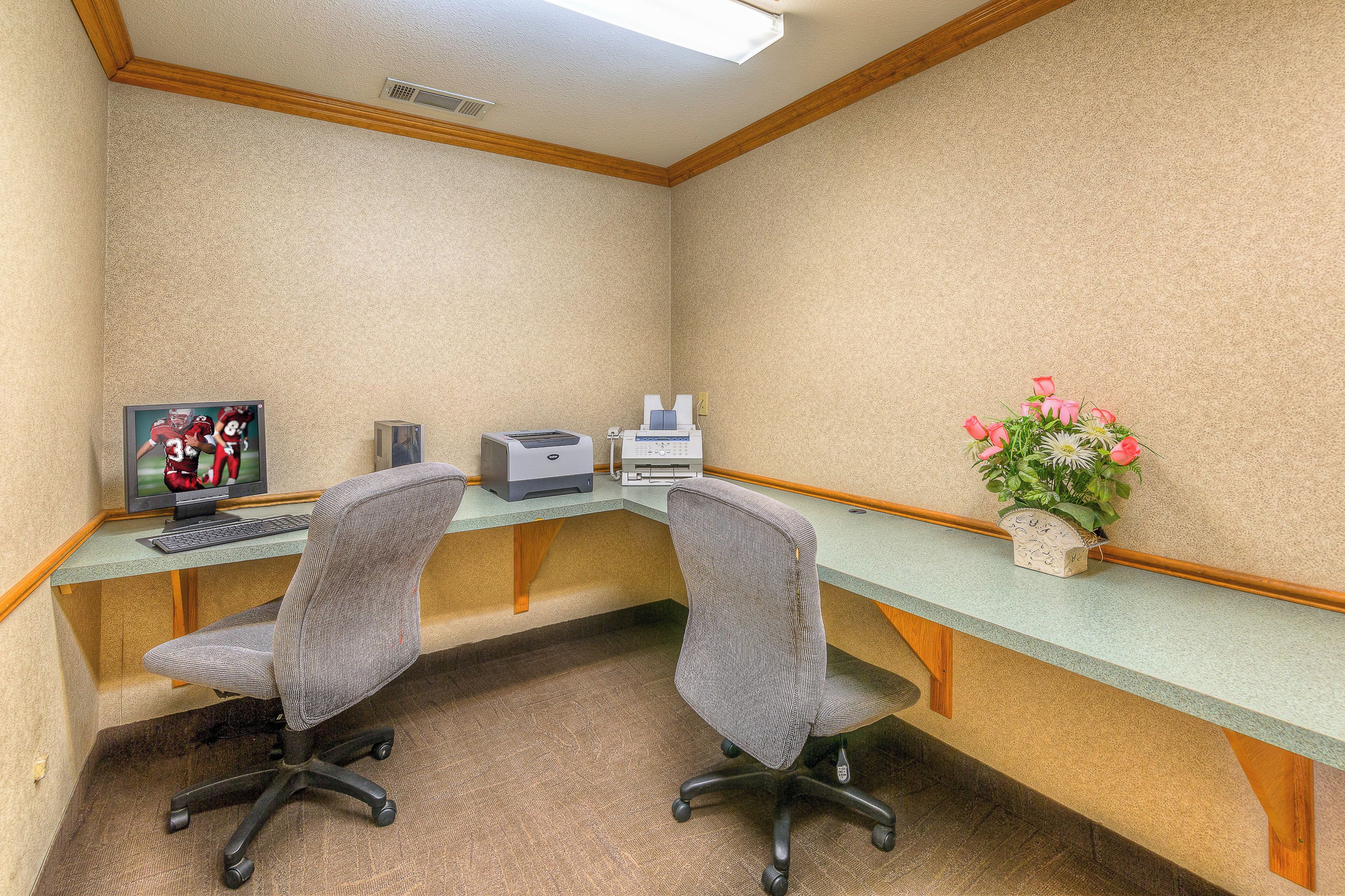 A corner office with two chairs, a desk, a printer, a computer monitor, and a vase with flowers.