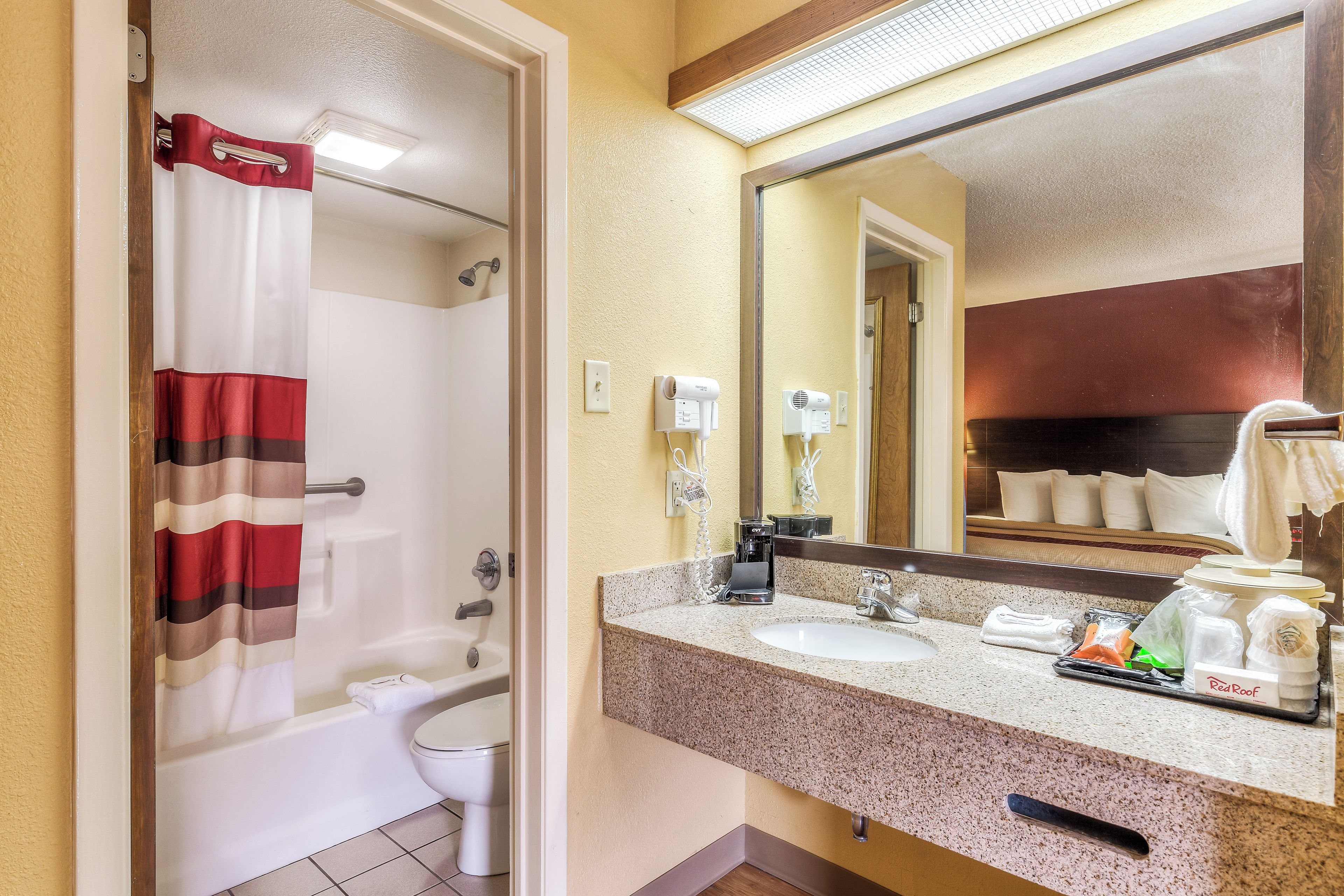 A hotel bathroom with a shower, toilet, sink, and a mirror reflecting a bed with pillows.