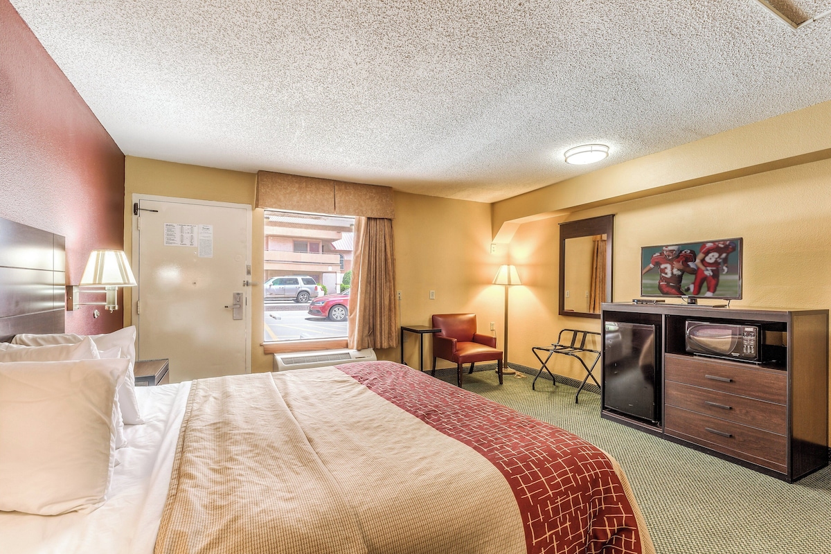 A hotel room with a bed, a desk with a microwave, a chair, a lamp, and a window with a view of the street.