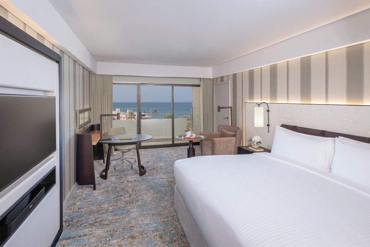 Habitación de hotel con una cama grande, un escritorio con silla, televisor y vista al océano.