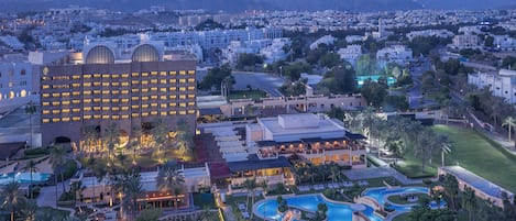Un hotel con un edificio grande, piscina y zona ajardinada con palmeras.