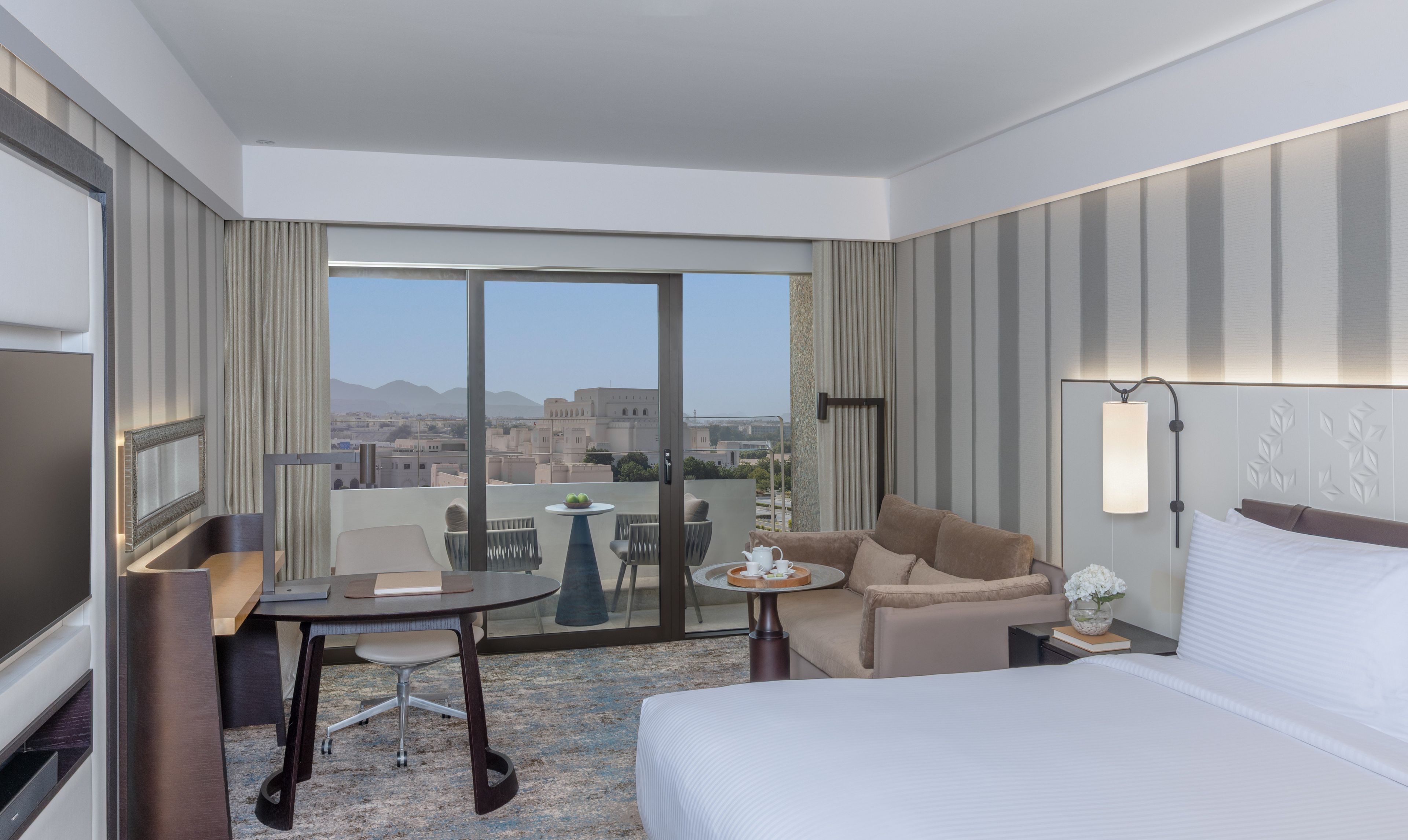 Habitación de hotel con cama, escritorio, silla, mesita, sofá y ventana grande con vistas a la ciudad.