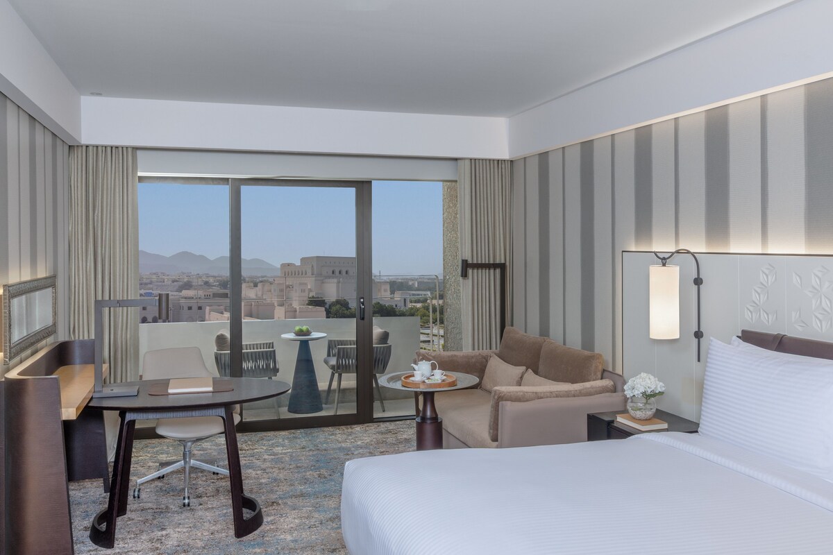 Habitación de hotel con cama, escritorio, silla, mesita, sofá y ventana grande con vistas a la ciudad.