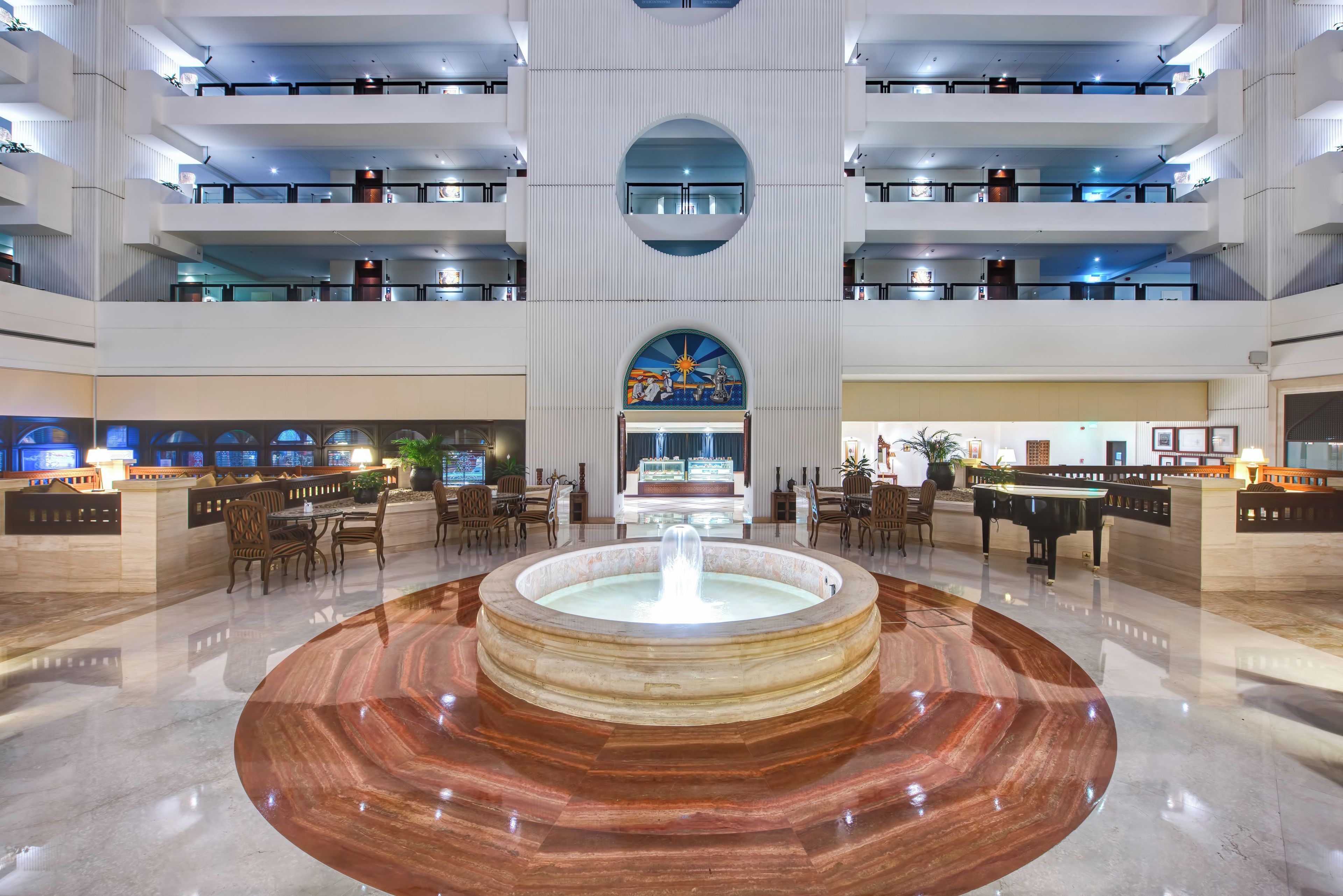 Un vestíbulo de hotel con una fuente central, varias zonas de comedor y un techo alto con iluminación moderna.