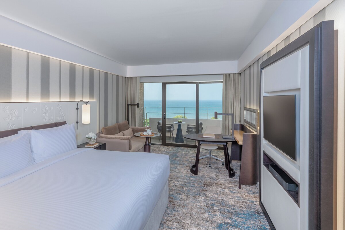 Habitación de hotel con una cama grande, un escritorio con silla, televisor y balcón con vista al océano.