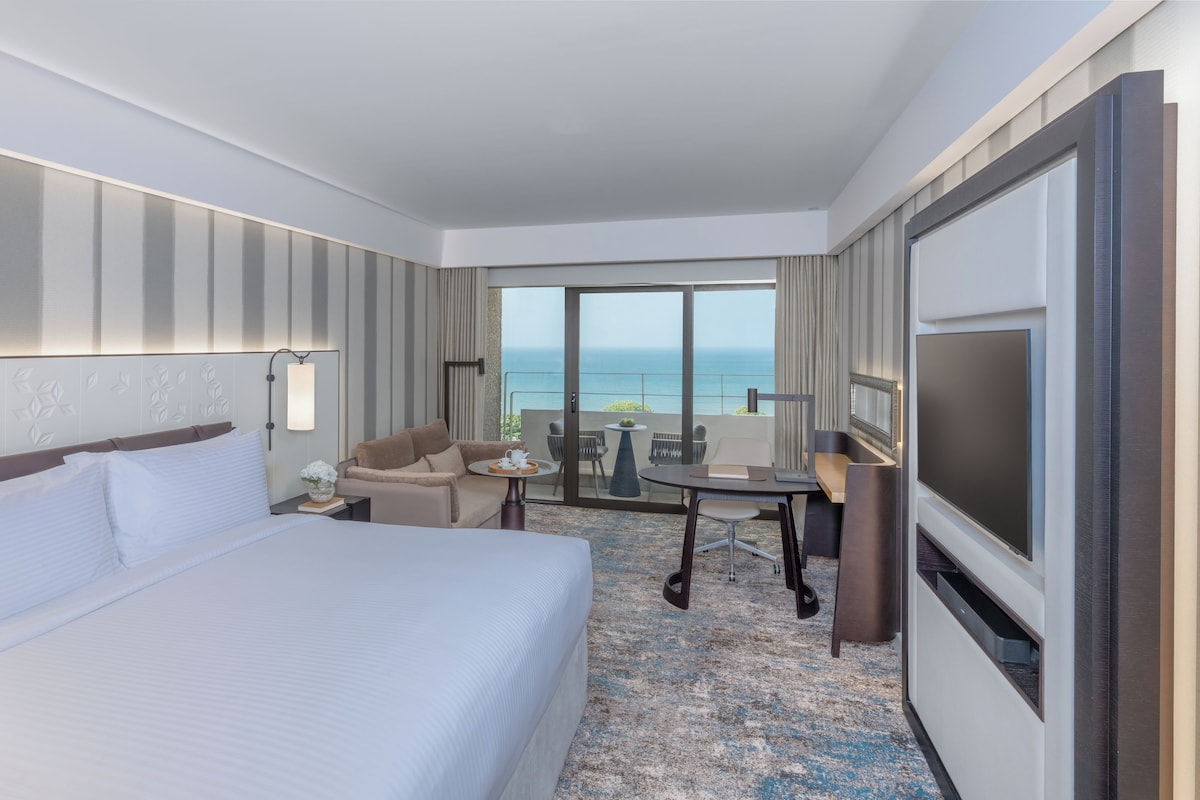 Habitación de hotel con una cama grande, un escritorio con silla, televisor y balcón con vista al océano.