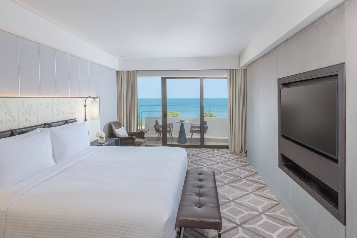 Habitación de hotel con una cama grande, televisión, banco, pequeña mesa con sillas y balcón con vista al océano.