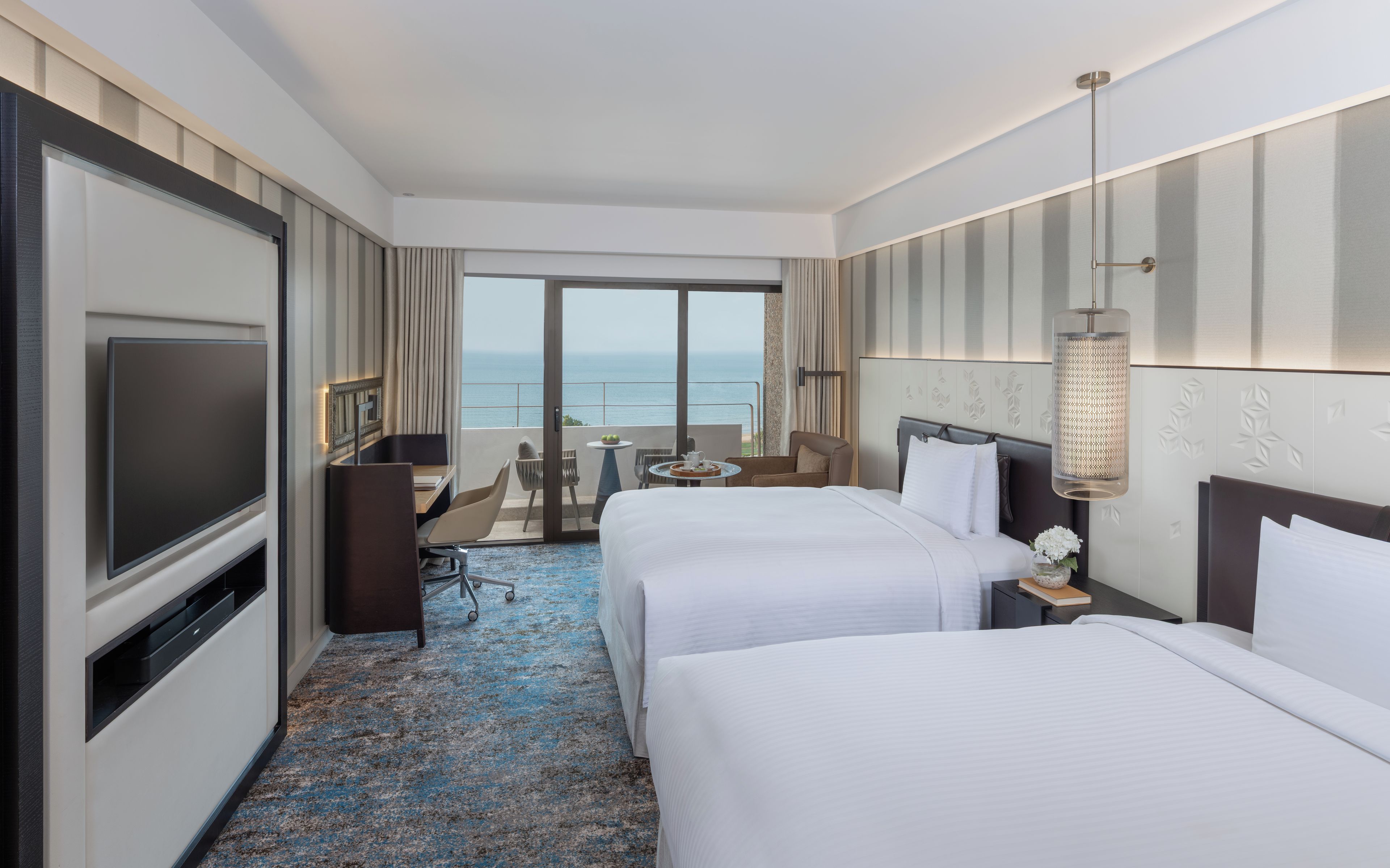 Habitación de hotel con dos camas, televisión, escritorio y balcón con vista al mar.