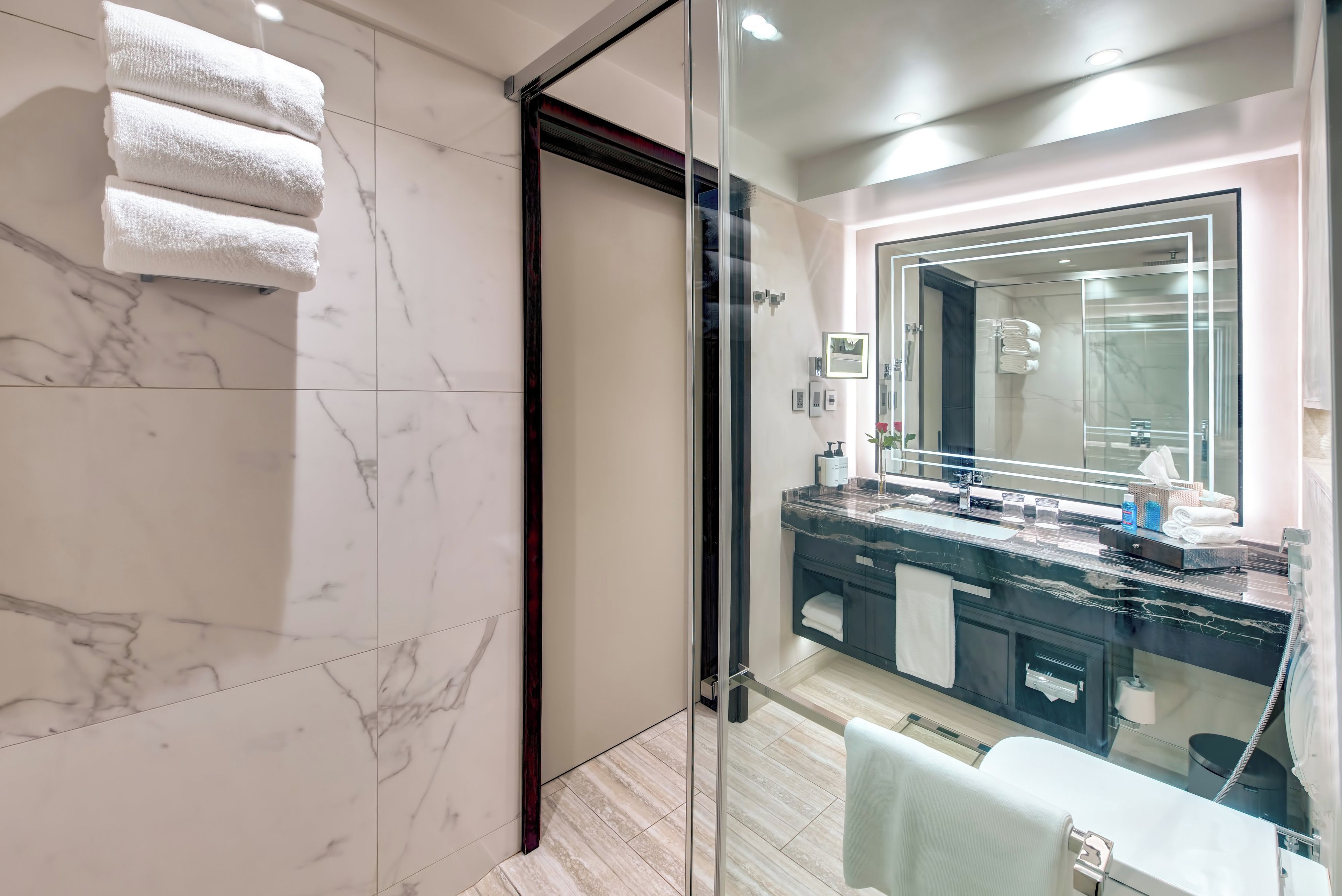 A modern bathroom with a glass shower enclosure, a marble-tiled wall, and a vanity with a large mirror.