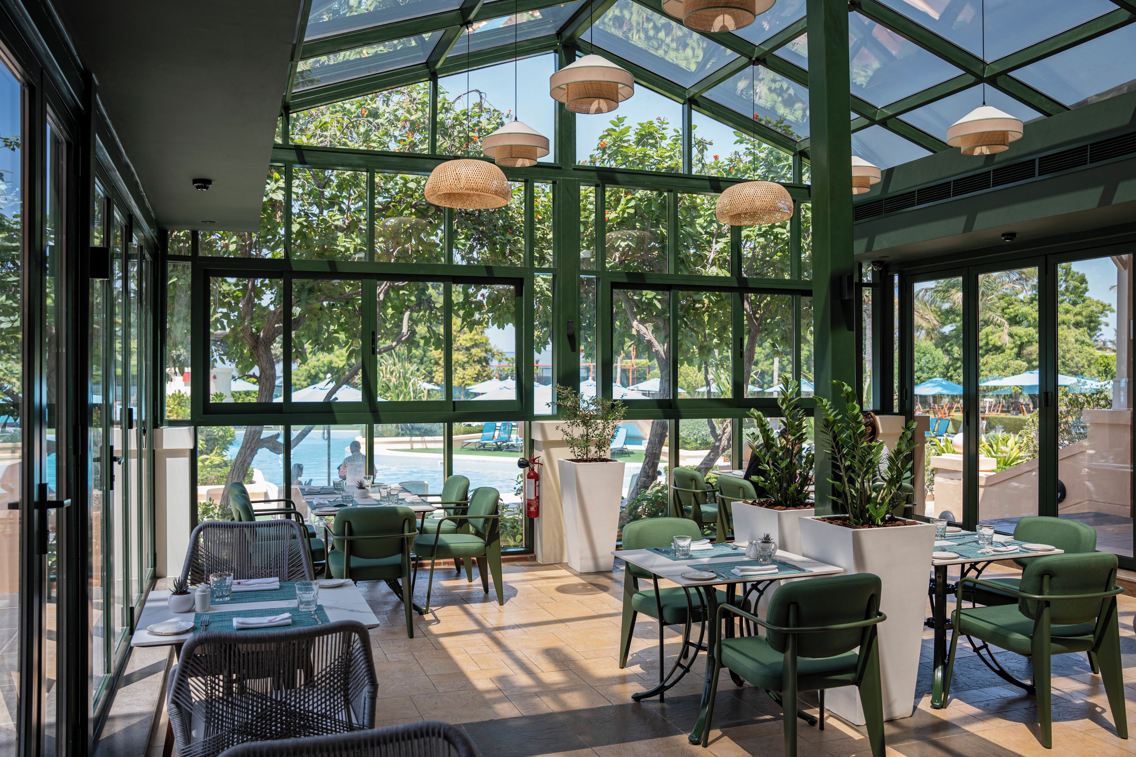 A restaurant with green chairs and tables, large windows, and hanging wicker lamps.
