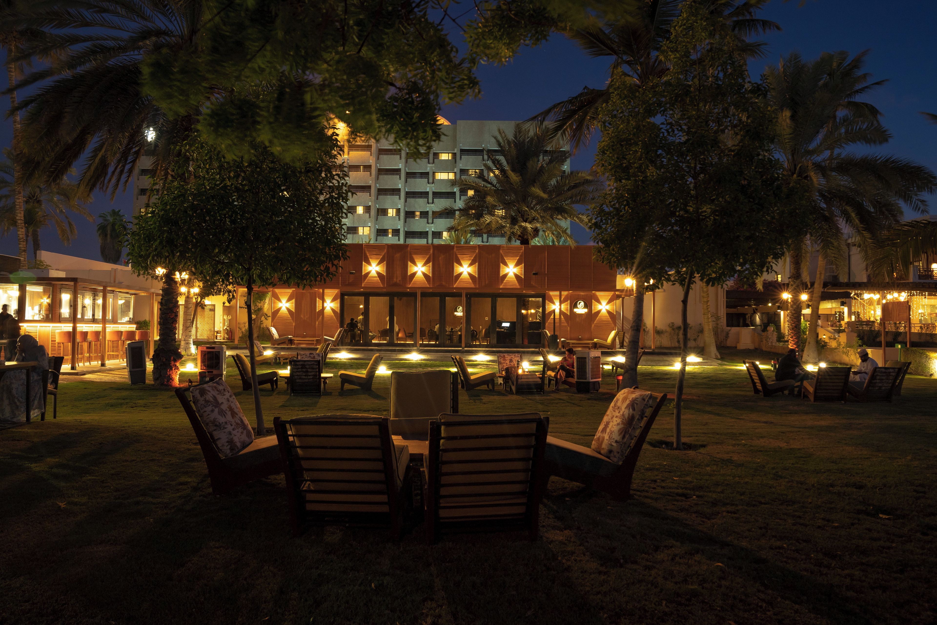 Un área de descanso al aire libre con asientos y una piscina, rodeada de palmeras y un edificio con ventanas iluminadas.
