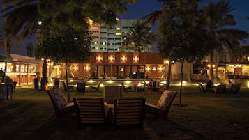 Un área de descanso al aire libre con asientos y una piscina, rodeada de palmeras y un edificio con ventanas iluminadas.