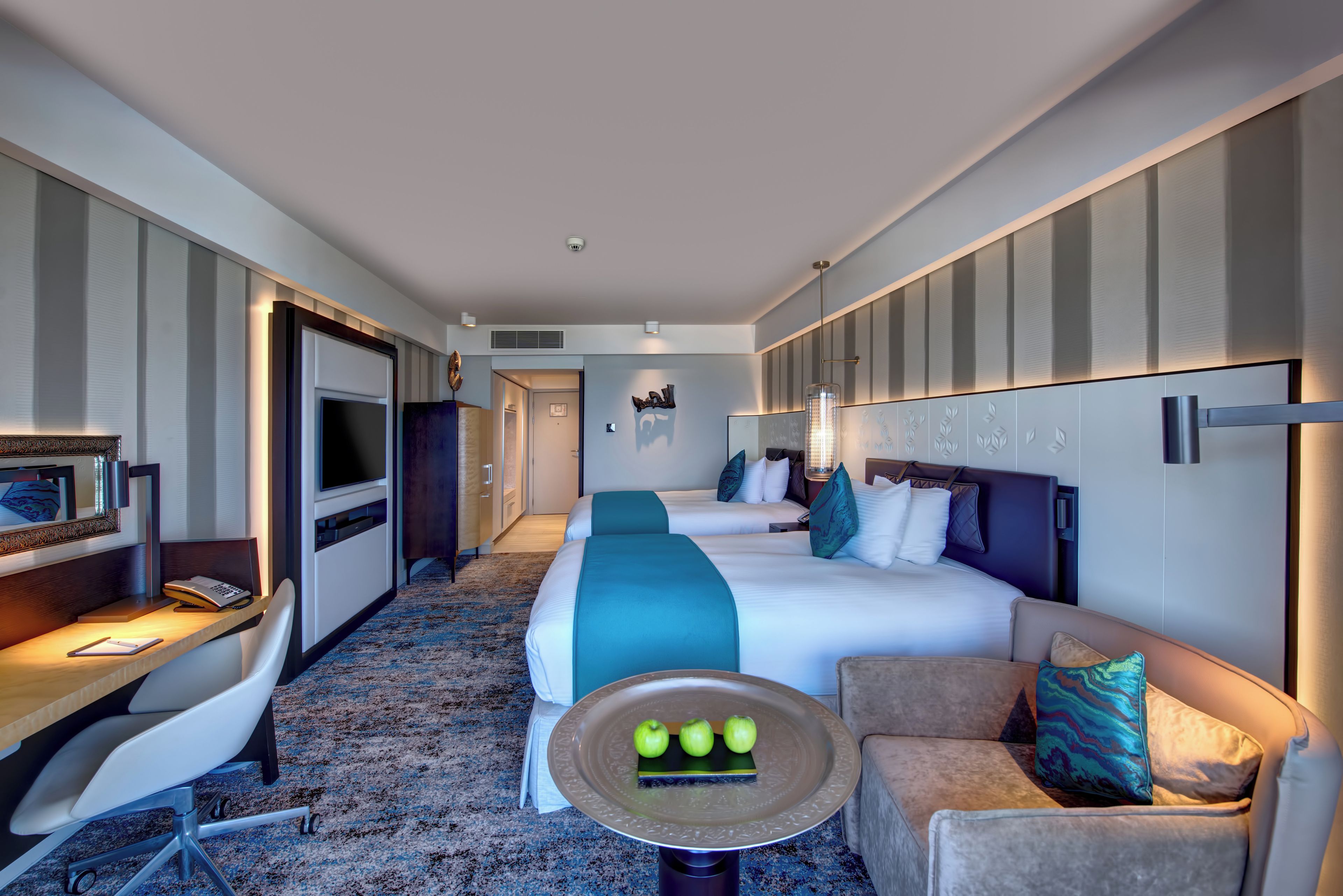 Een hotelkamer met een bed, een bureau met een computer, een stoel, een kleintje tafeltje met appels en een televisie.
