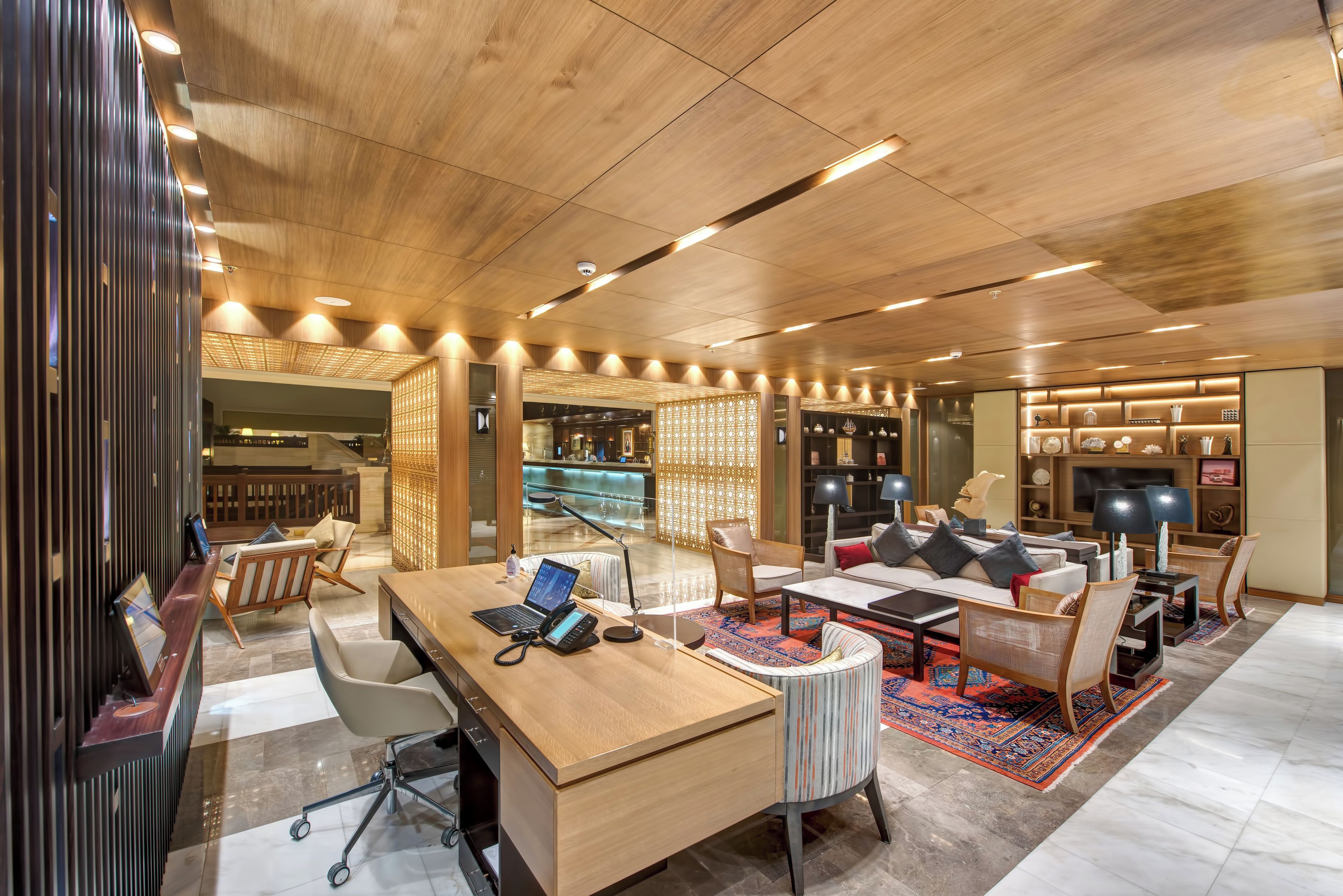 A modern hotel lobby with a wooden reception desk, seating area, and a TV.