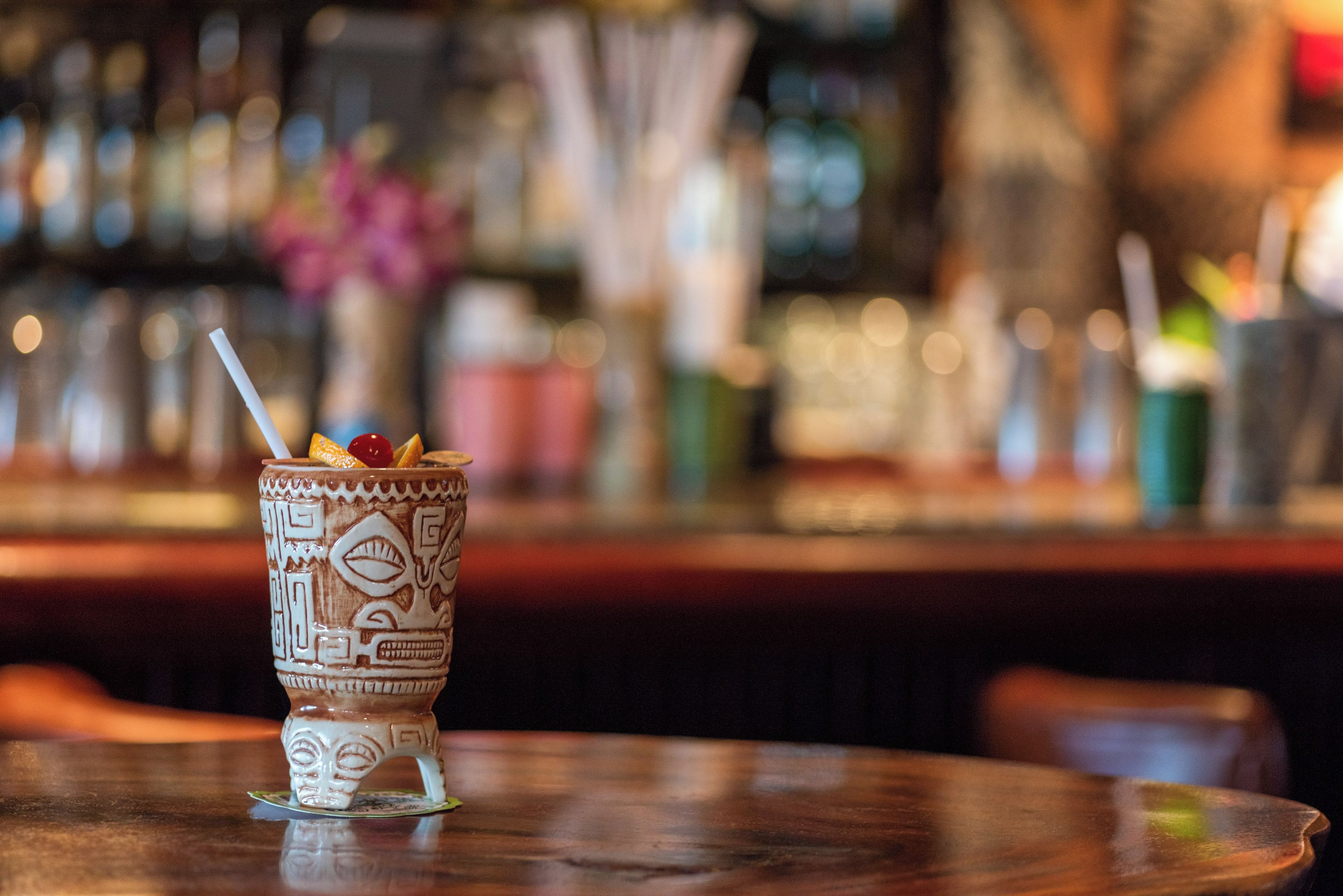 Un ambiente de bar con un vaso decorativo que contiene una bebida y una pajita, una barra y un fondo borroso con botellas y vasos.